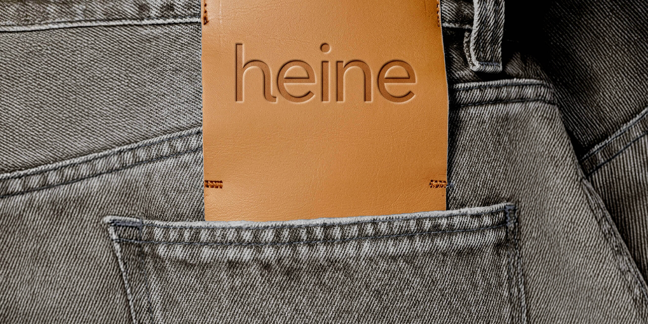 Nahaufnahme der Gesäßtasche einer grauen Jeans. Aus der Tasche ragt ein braunes Leder-Etikett mit dem eingeprägten Schriftzug 'heine'.