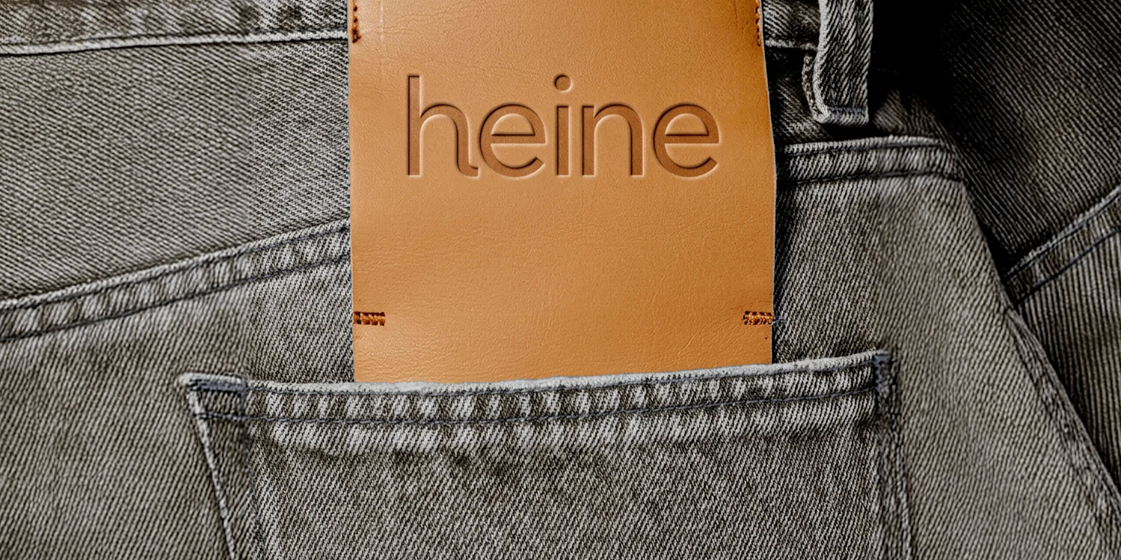 Nahaufnahme der Gesäßtasche einer grauen Jeans. Aus der Tasche ragt ein braunes Leder-Etikett mit dem eingeprägten Schriftzug 'heine'.