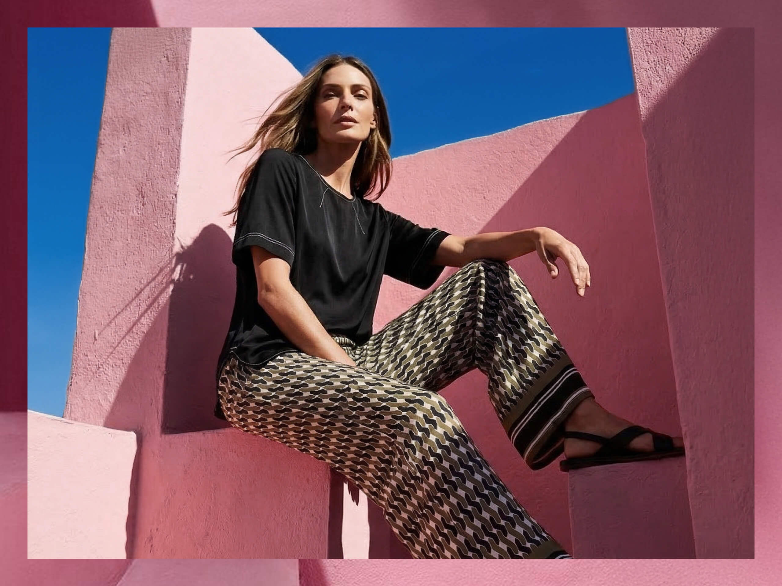 Een model zit op een roze trap en draagt een zwart T-shirt met korte mouwen en wijde, gemêleerde broek in zwart, wit en beige.