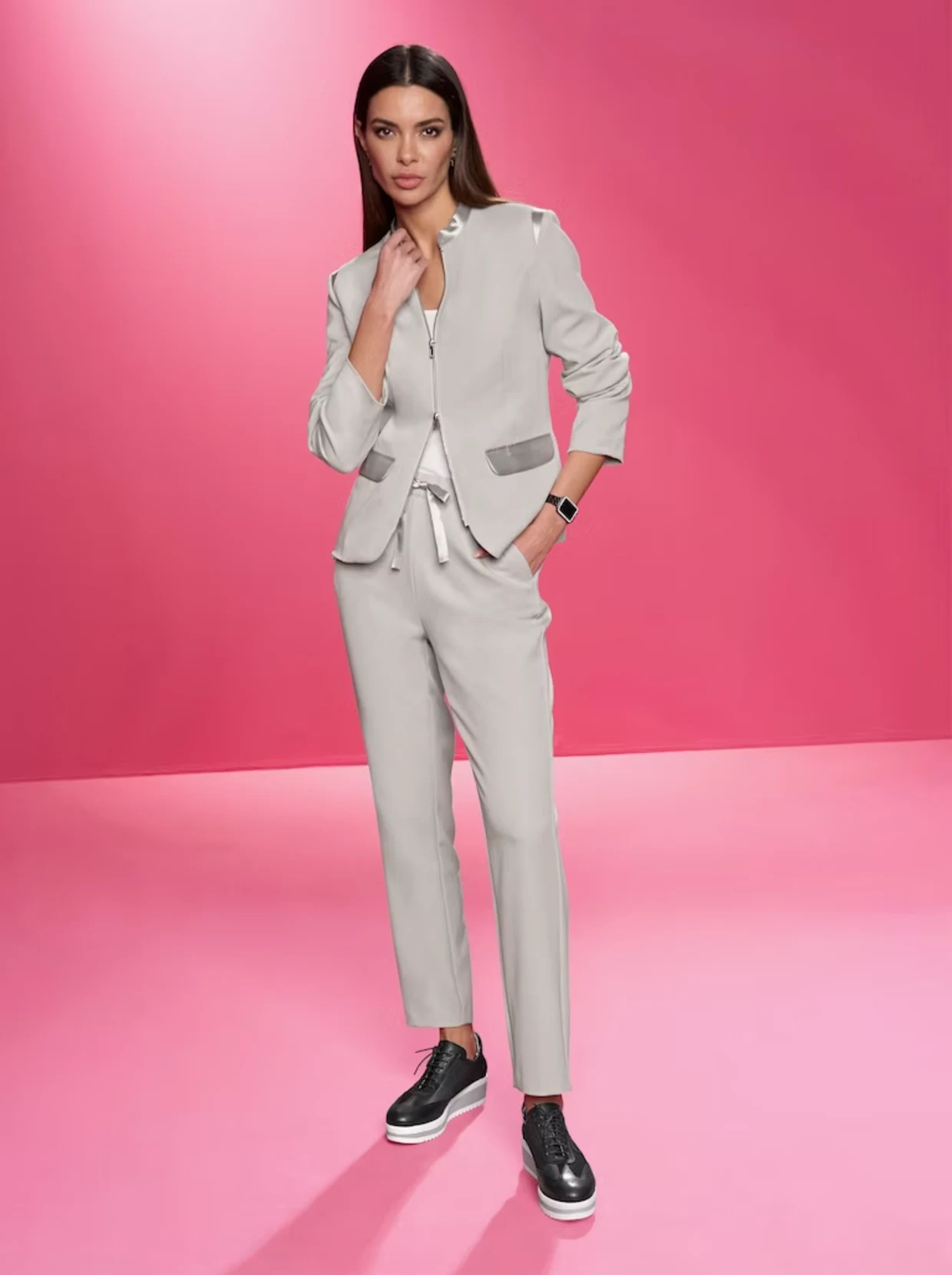 Ein Model präsentiert einen lässigen grauen Hosenanzug mit einem Reißverschluss und einem Gürtel, kombiniert mit sportlichen schwarzen Sneakers. Der Hintergrund in kräftigem Pink.