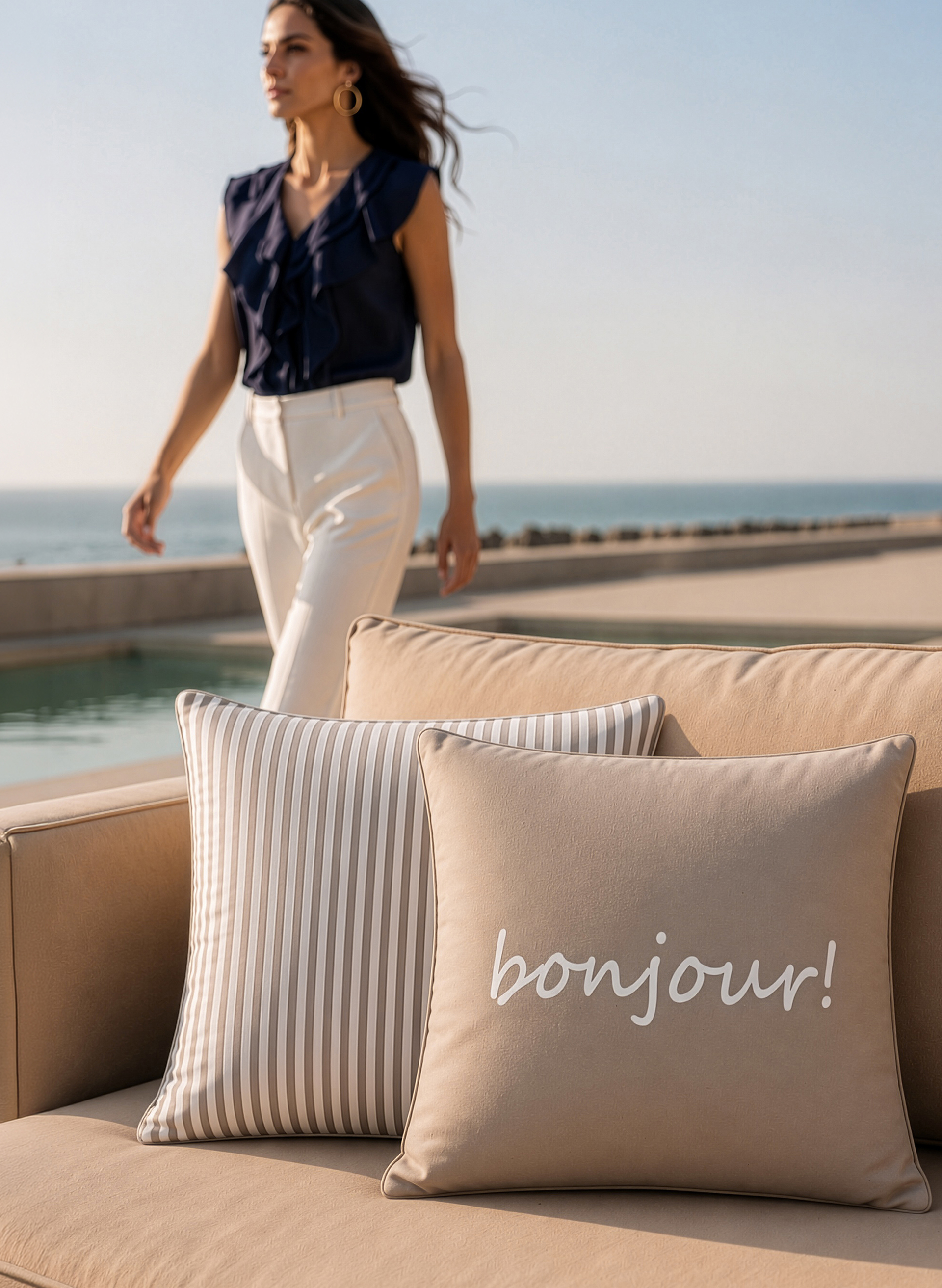 Zwei Kissen liegen auf einem beigen Sofa, eines gestreift, das andere mit dem Schriftzug 'bonjour!'. Im Hintergrund geht ein Model mit dunkelblauer Bluse und heller Hose am Wasser entlang.