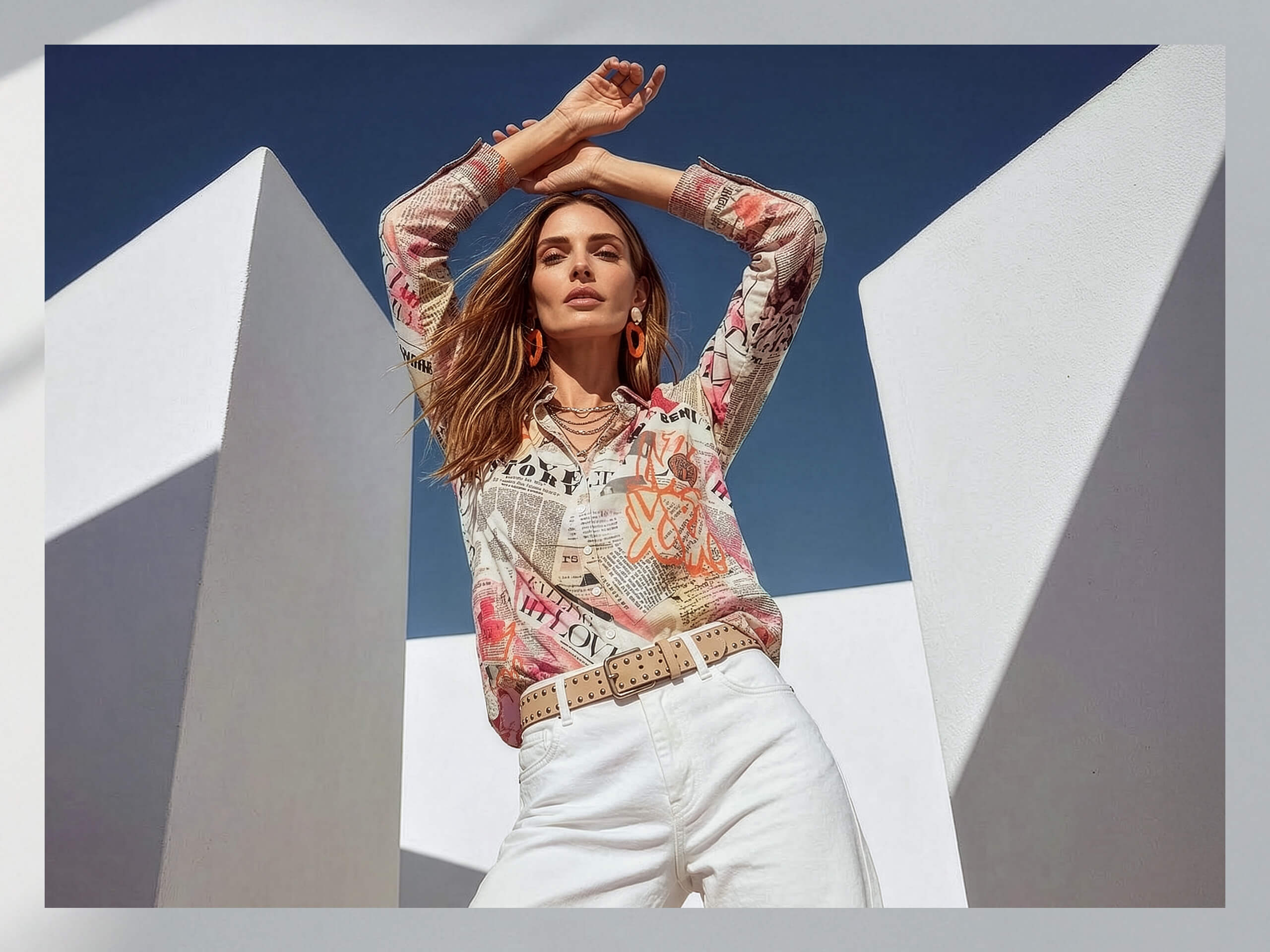 Een model staat voor een wit, modern gebouw, draagt een kleurrijk blouse met tekstpatronen en witte broek, de armen zijn boven het hoofd gekruist.