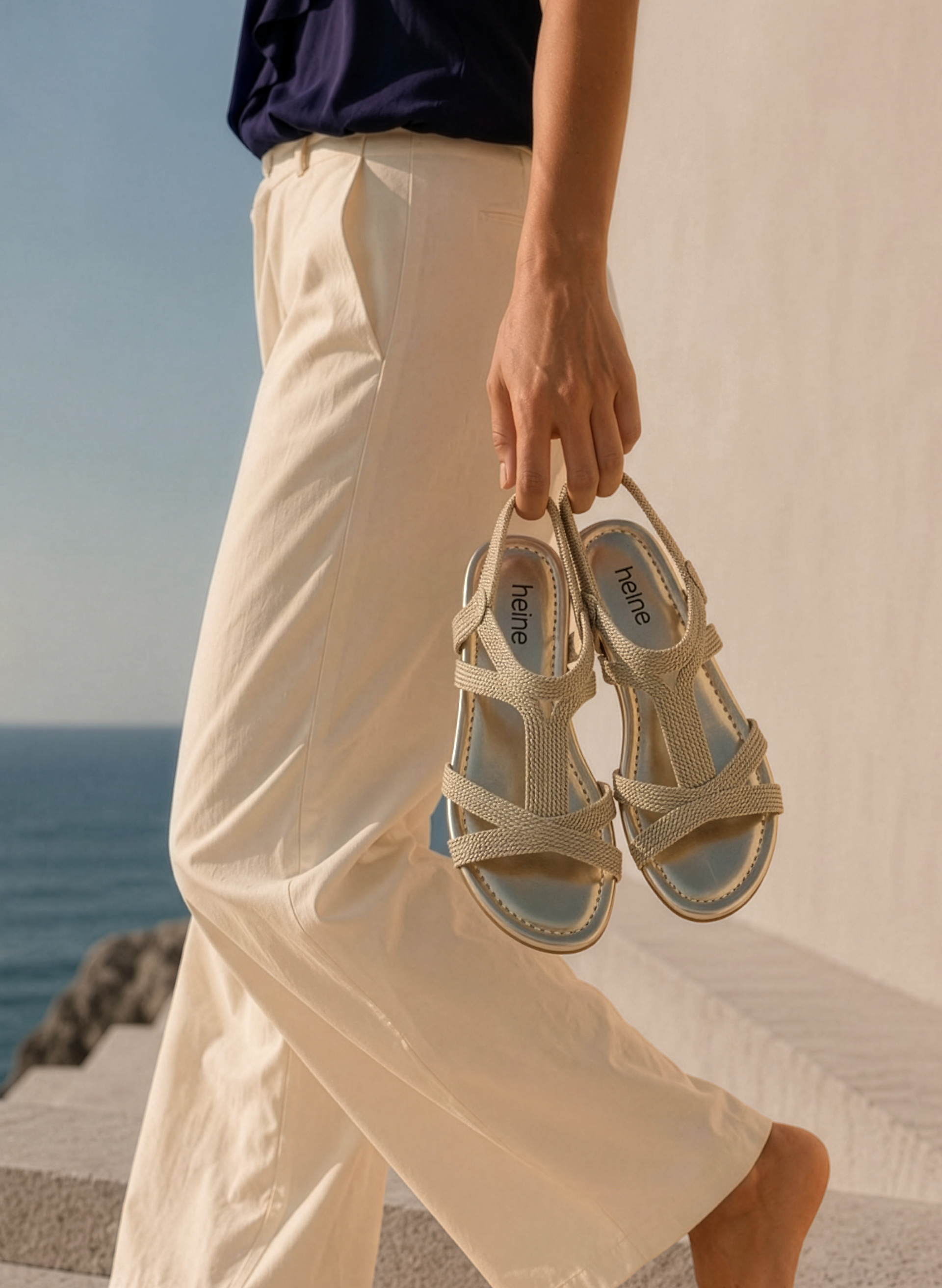 Un modèle tient une paire de sandales beige, porte un pantalon clair. En arrière-plan, on voit la mer.