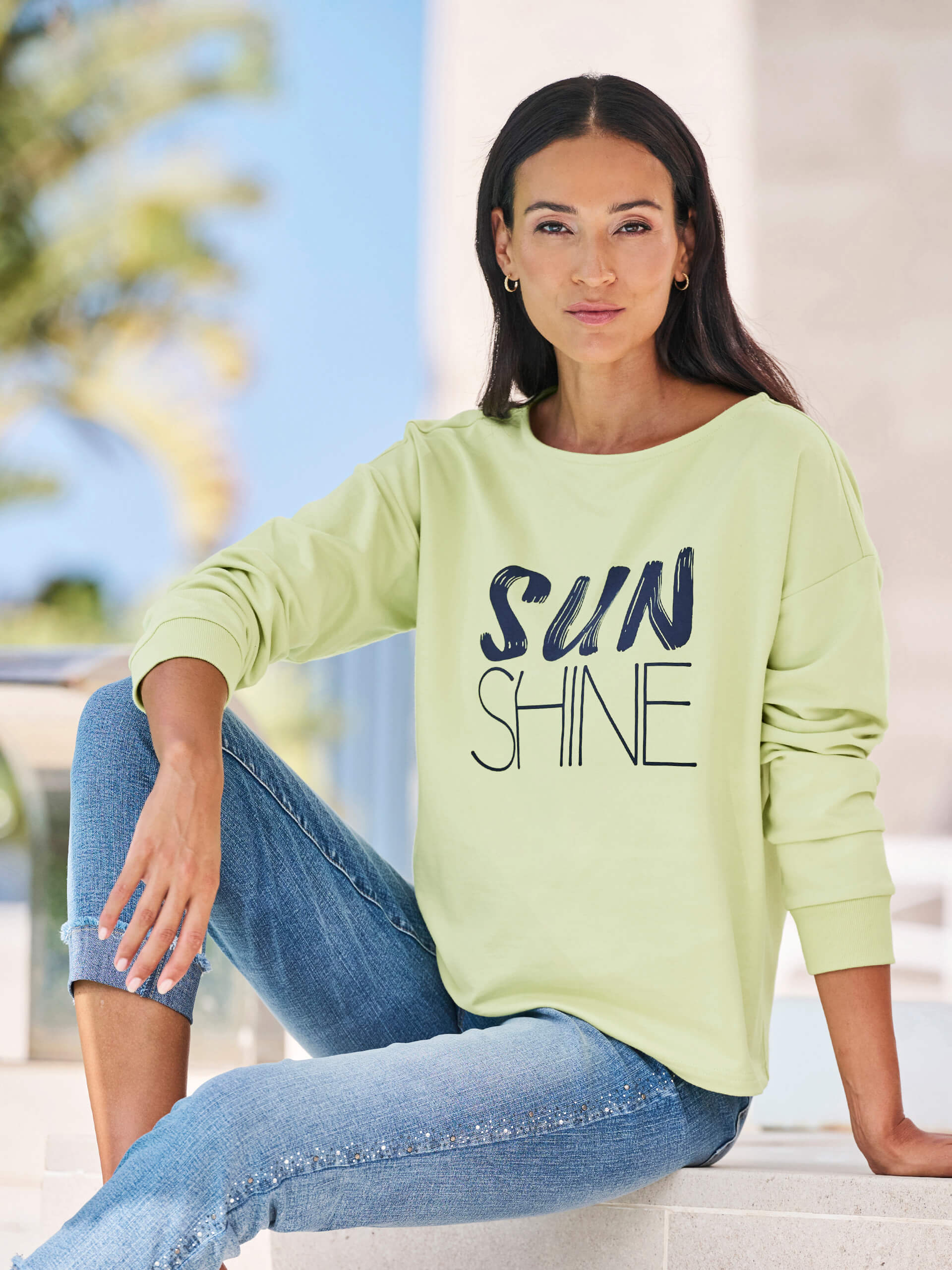 Ein Model sitzt entspannt und trägt ein hellgrünes Sweatshirt mit dem Schriftzug 'SUNSHINE' und blaue Jeans.