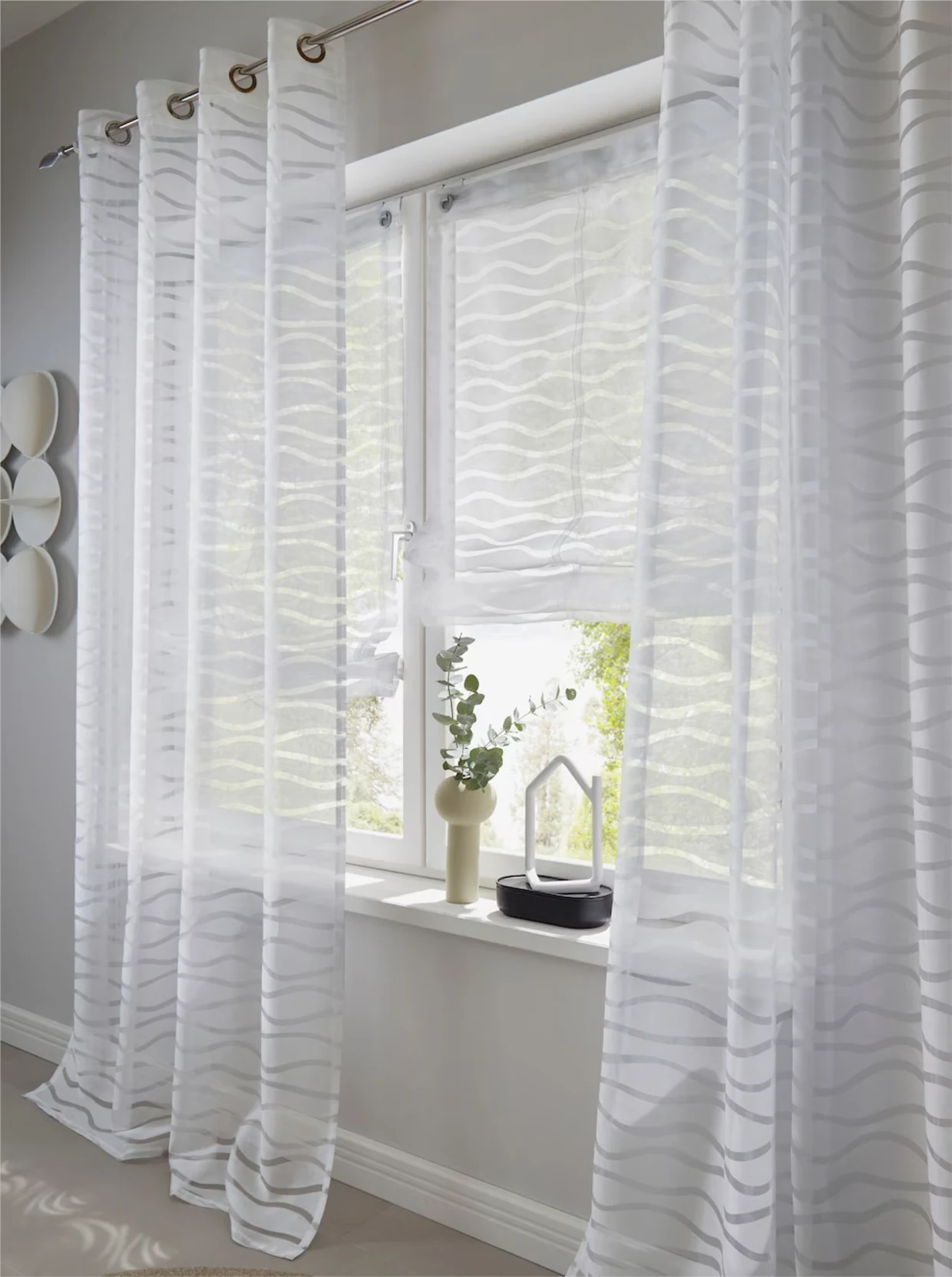 Twee transparante gordijnen met een golvend patroon hangen aan een raam, dat wordt aangevuld door een rolgordijn met een soortgelijk ontwerp. Twee transparante gordijnen met een golvend patroon hangen aan een raam, dat wordt aangevuld door een rolgordijn met een soortgelijk ontwerp.