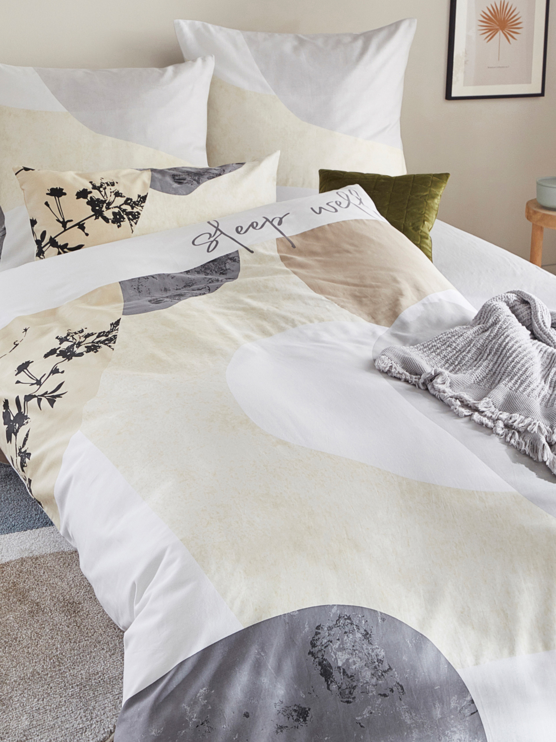 Ein Bett mit hell gemusterter Bettwäsche in Beige, Grau und Weiß, darauf liegt eine graue gestrickte Decke, im Hintergrund Kissen und ein Bild an der Wand.