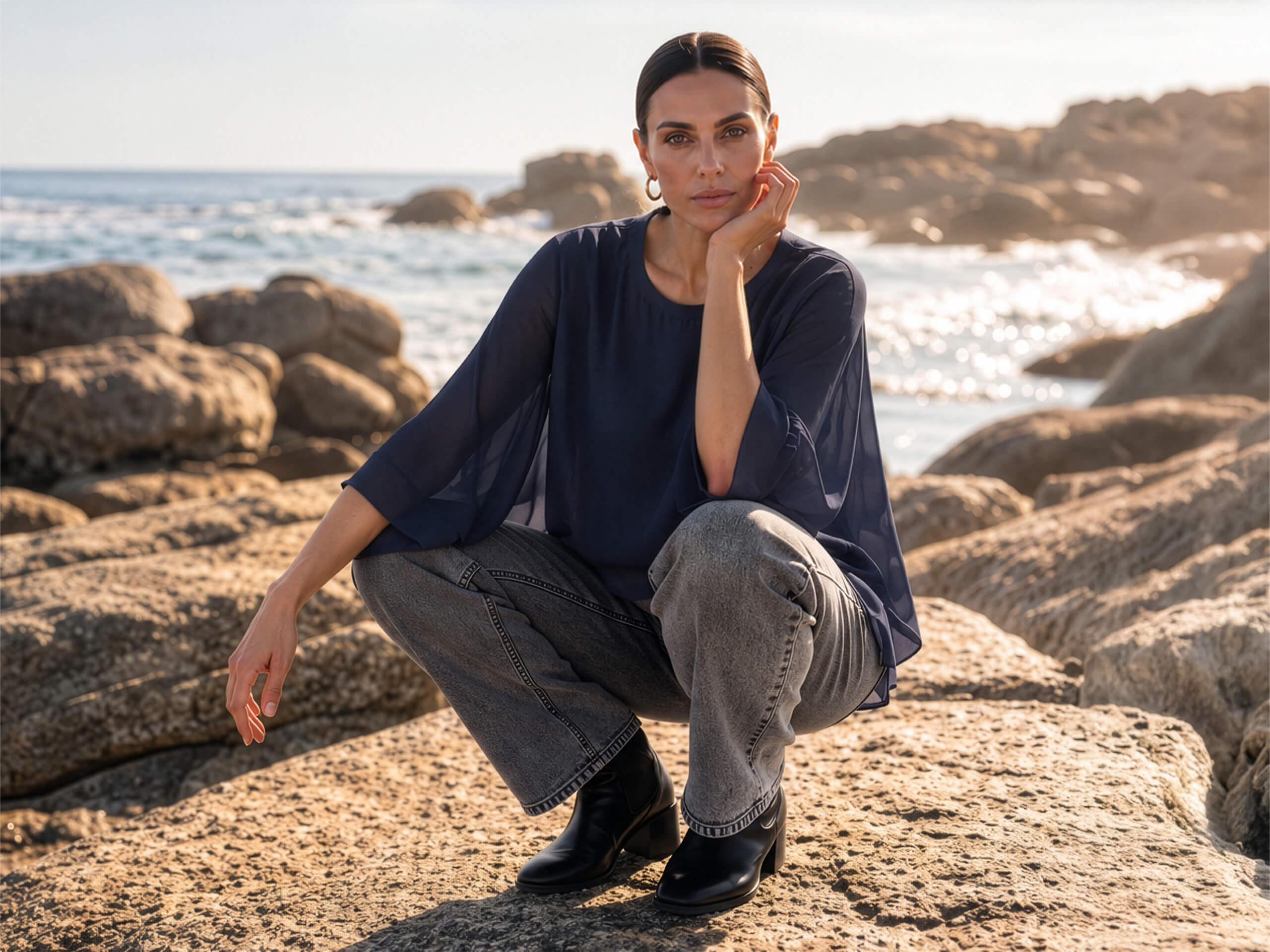 Un mannequin en blouse bleue et jean gris est accroupi sur des rochers sur la côte. Elle appuie son menton sur sa main et regarde l'appareil photo.