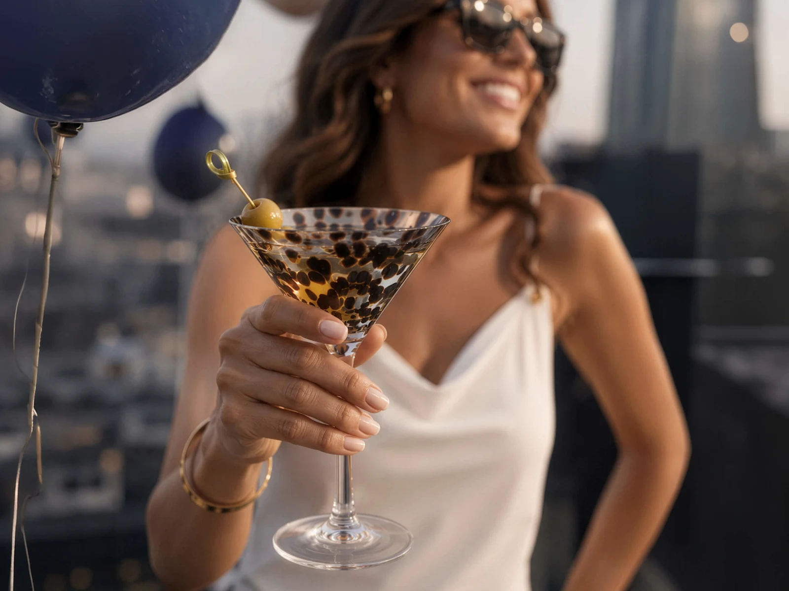 Ein Model mit Sonnenbrille und weißem Top hält ein Cocktailglas mit einem Oliven-Spieß, im Hintergrund unscharfe Ballons und Stadtansicht.