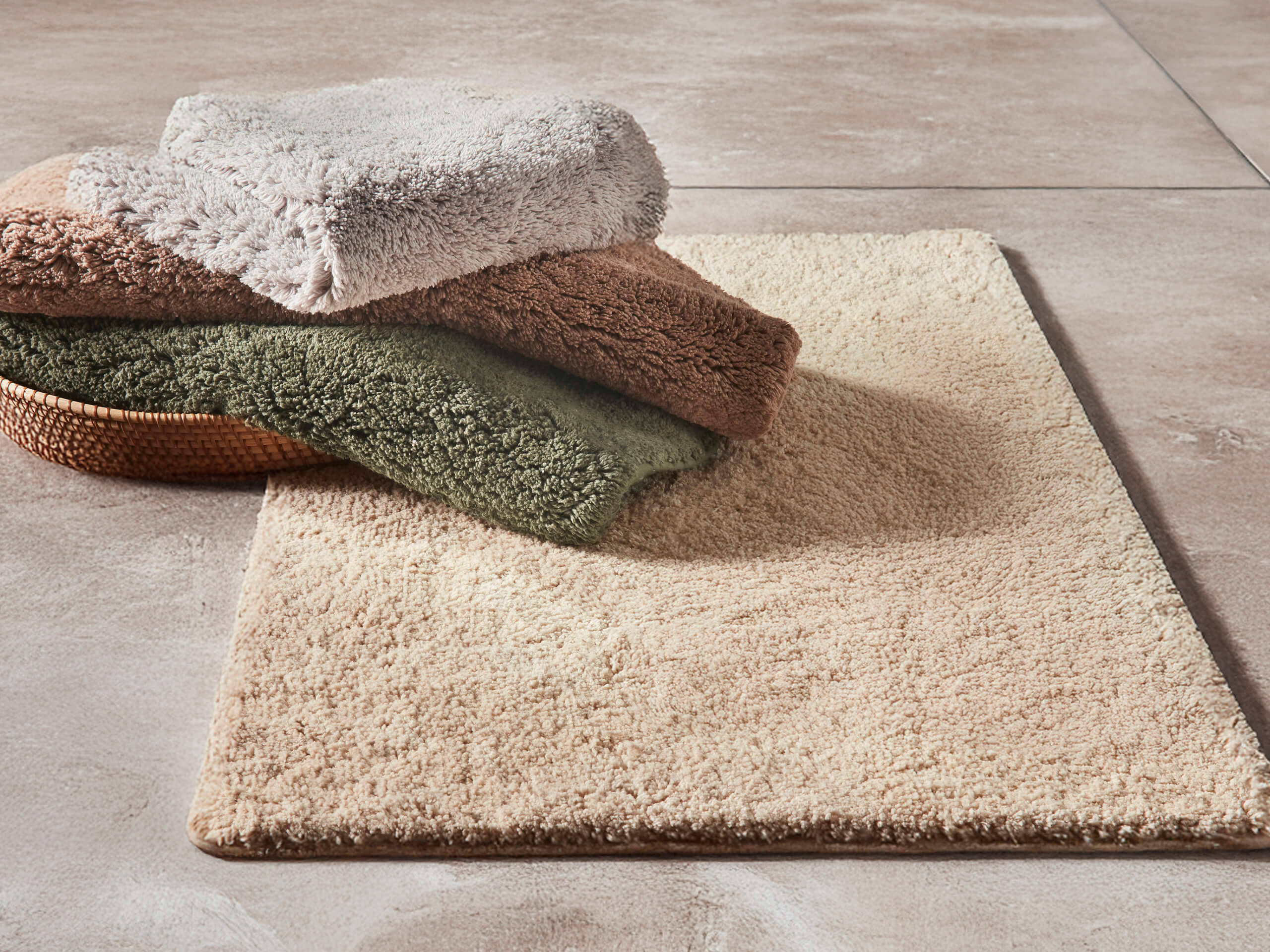 Auf einem beigen Badteppich liegen drei gefaltete Handtücher in Beige, Braun und Grün. Der Boden ist gefliest.
