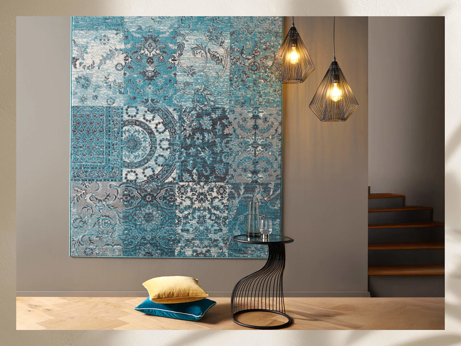 Tapis mural avec motif patchwork bleu-gris, à côté deux lampes suspendues en fil métallique, petite table ronde avec bouteille en verre et deux coussins au sol.