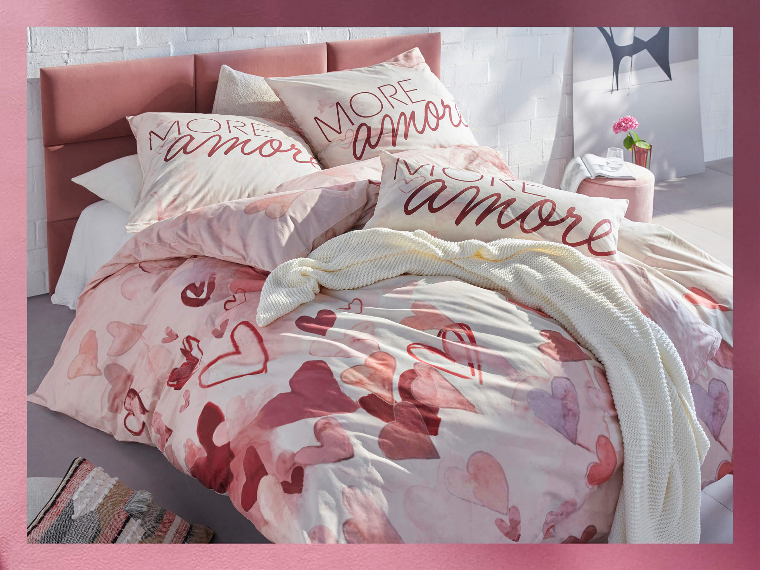 Een bed met roze beddengoed met hartmotief en kussens met de tekst 'More amor'. Daarnaast twee tapijten in roze en beige met een reliëf bloemmotief.