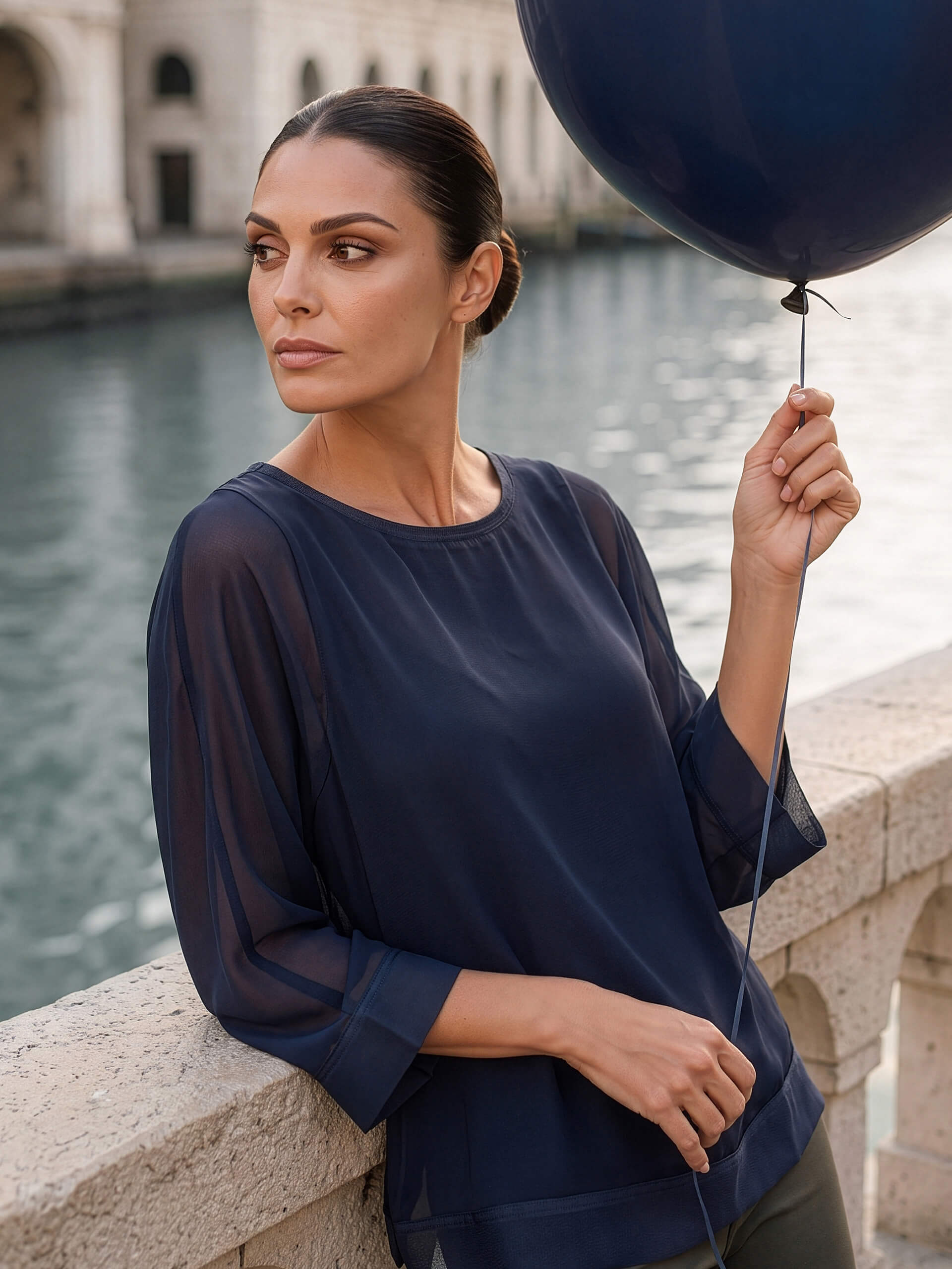 Ein Model steht an einem Kanal, trägt eine dunkelblaue Bluse und hält einen dunkelblauen Ballon.