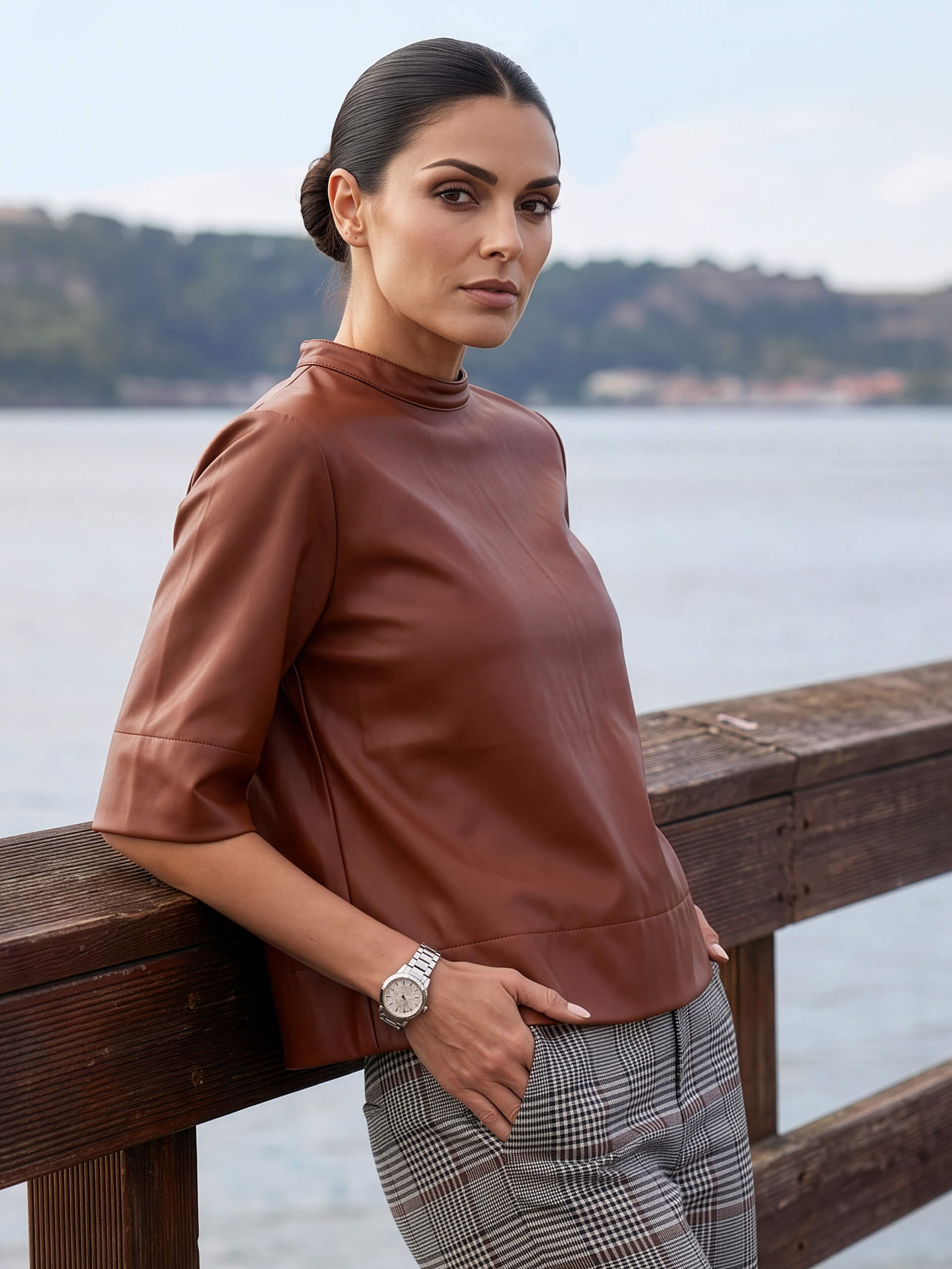 Ein Model in einem braunen Oberteil lehnt an einem Holzgeländer am Wasser.