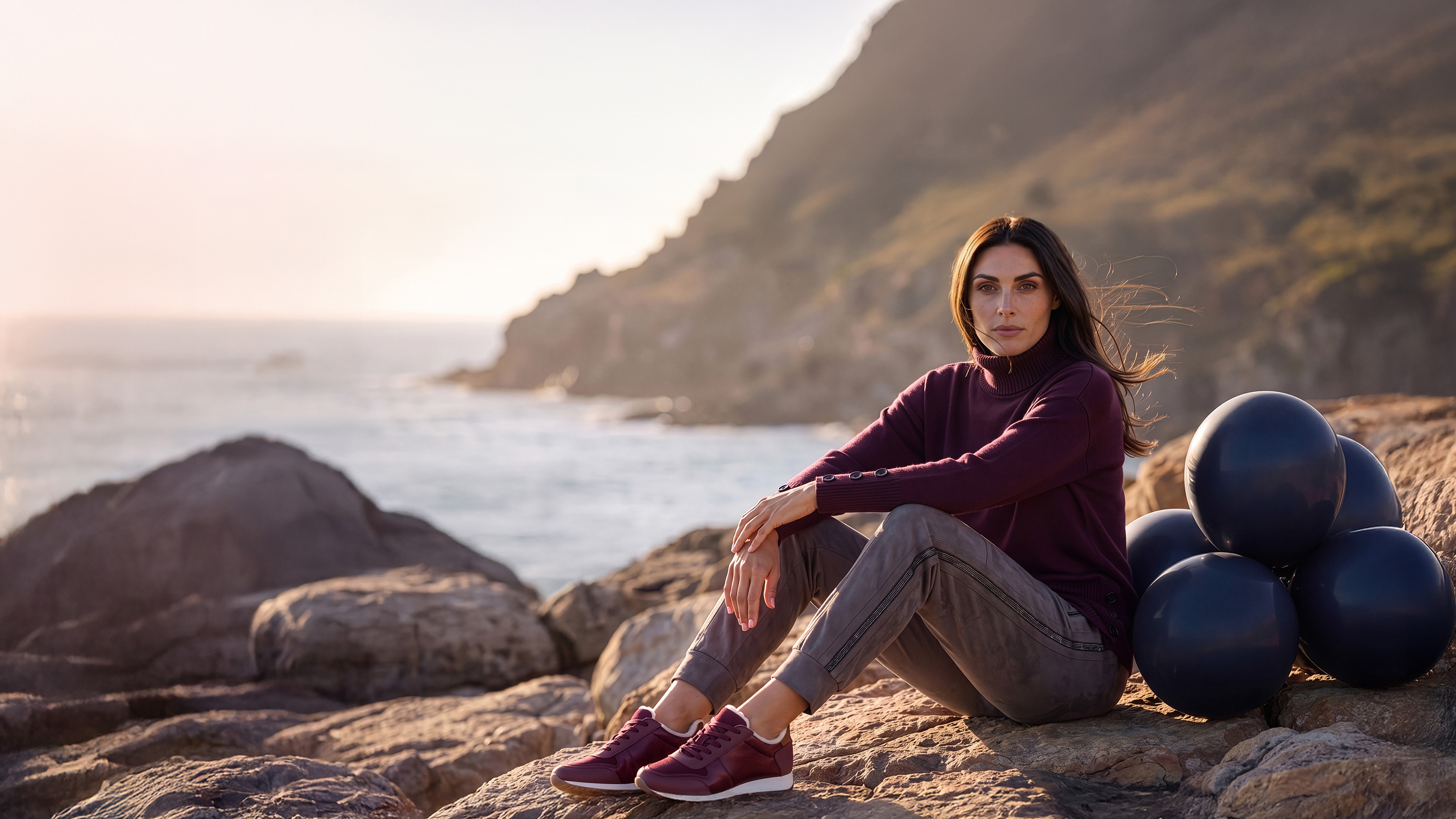 Een model zit op een rots. Ze draagt een bordeauxrode coltrui, een grijze broek en bordeauxrode sneakers