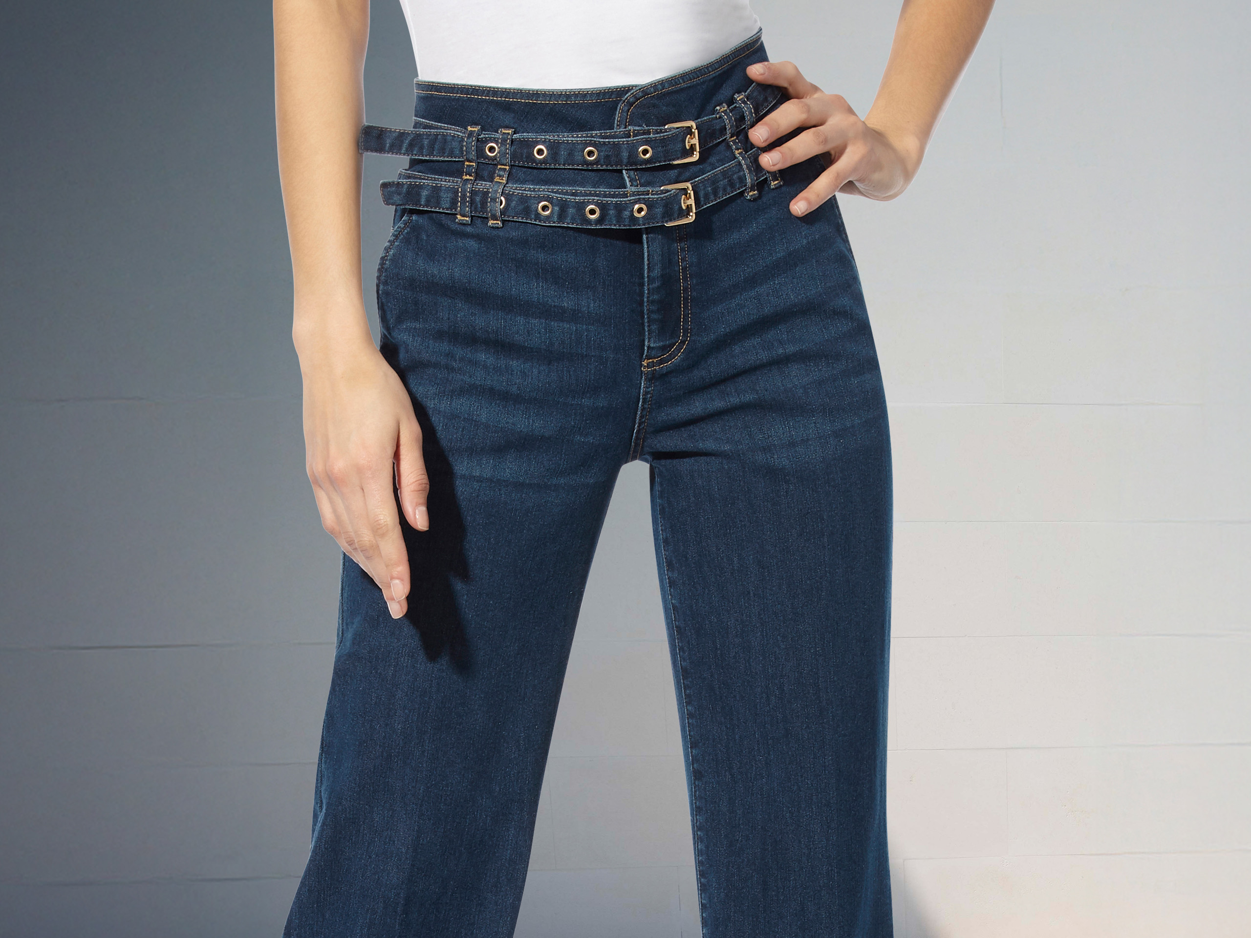 Eine Hose aus dunklem Denim mit weitem Bein, hohem Bund und doppeltem Gürtel mit Schnallen, getragen mit weißem Oberteil.