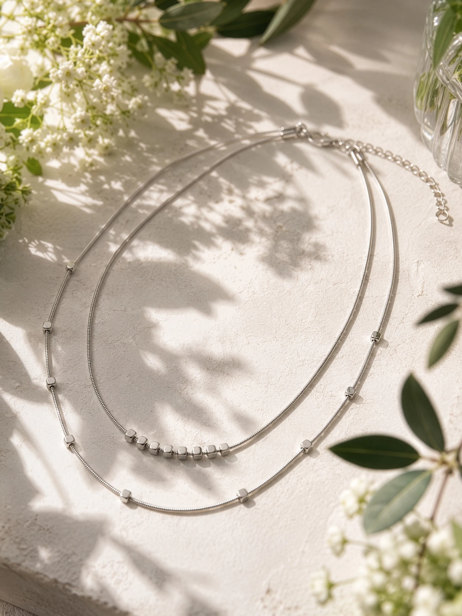 Deux fines chaînes en argent avec de petites perles reposent sur un fond de pierre claire, entourées de feuilles vertes et de fleurs blanches.