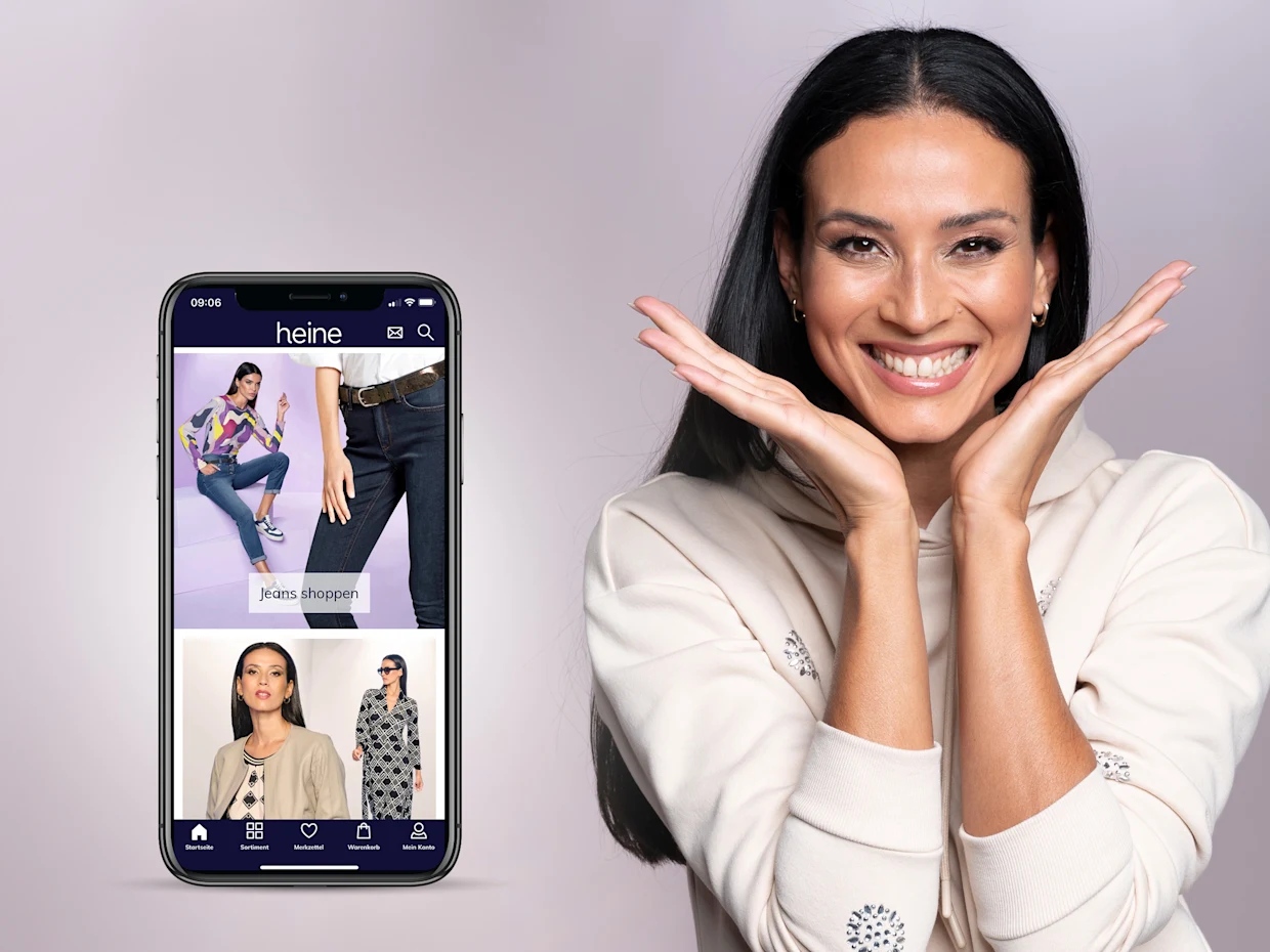 Smartphone mit geöffneter Heine-App, die Kleidung und Modeangebote zeigt, daneben eine Person in einem cremefarbenen Hoodie mit ausgestreckten Händen vor neutralem Hintergrund. Smartphone mit geöffneter Heine-App, die Kleidung und Modeangebote zeigt, daneben eine Person in einem cremefarbenen Hoodie mit ausgestreckten Händen vor neutralem Hintergrund.