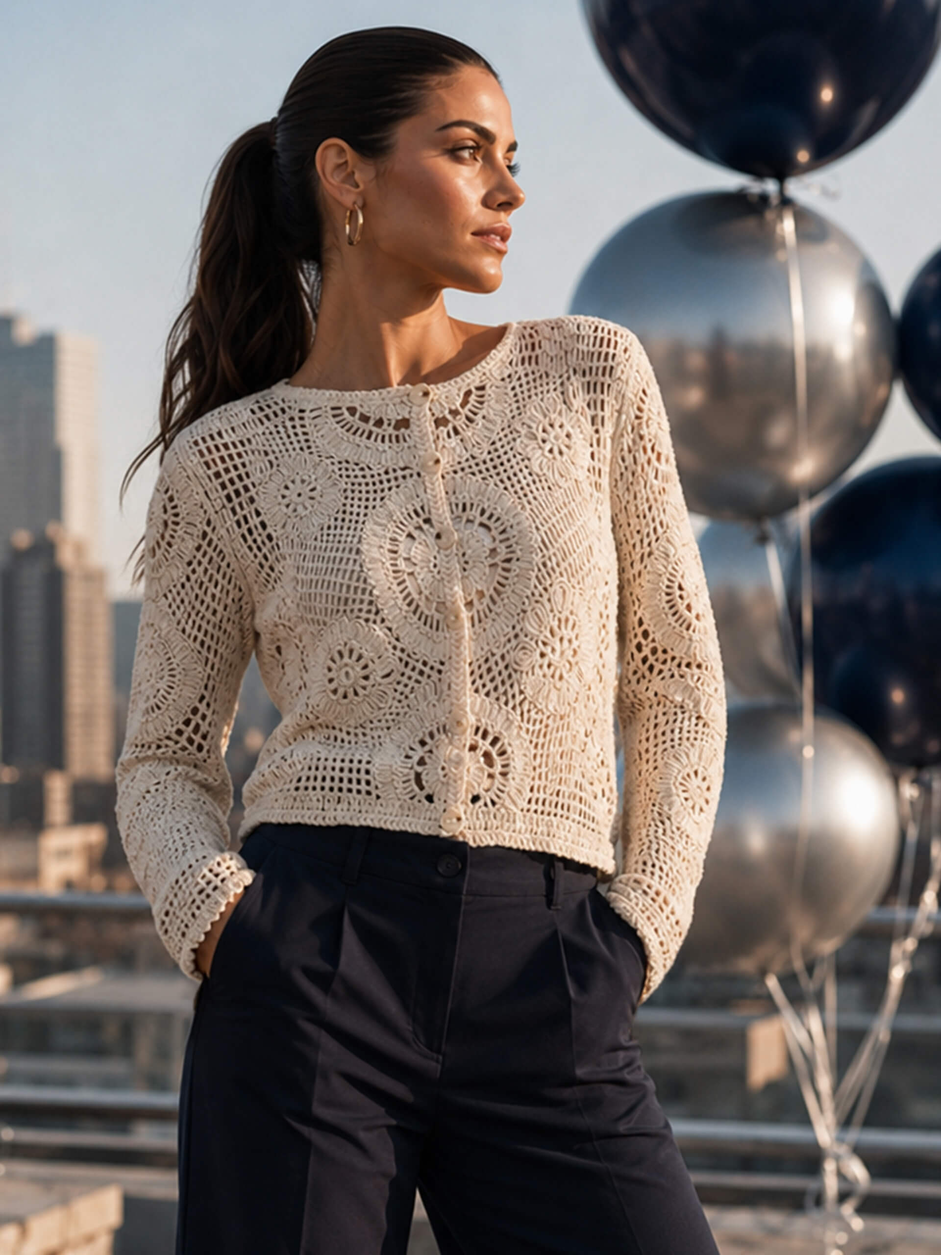 Un modèle porte un gilet beige en crochet avec des boutons, en arrière-plan une ville et des ballons argentés et bleus.