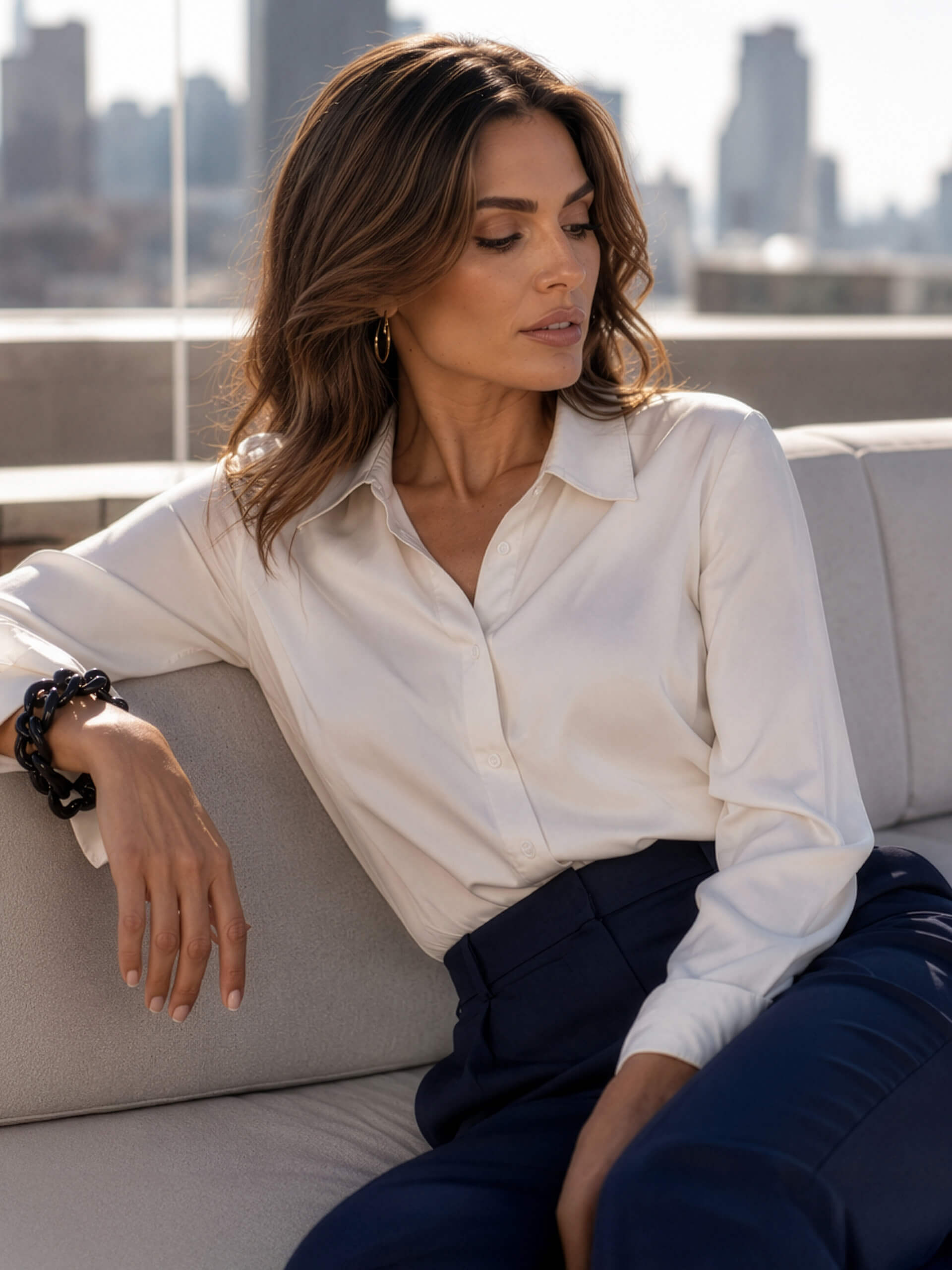 Een model zit op een lichte bank, draagt een witte blouse en donkerblauwe broek, op de achtergrond een vage stadsachtergrond.