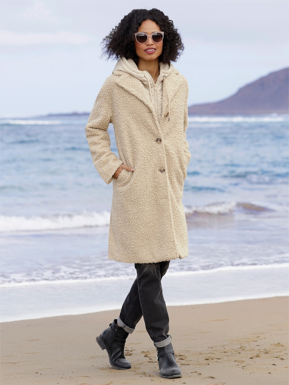 Model steht am Strand und trägt einen sandfarbenen Mantel mit herausnehmbarer Steppweste, Reverskragen, Knopfleiste und Taschen. 