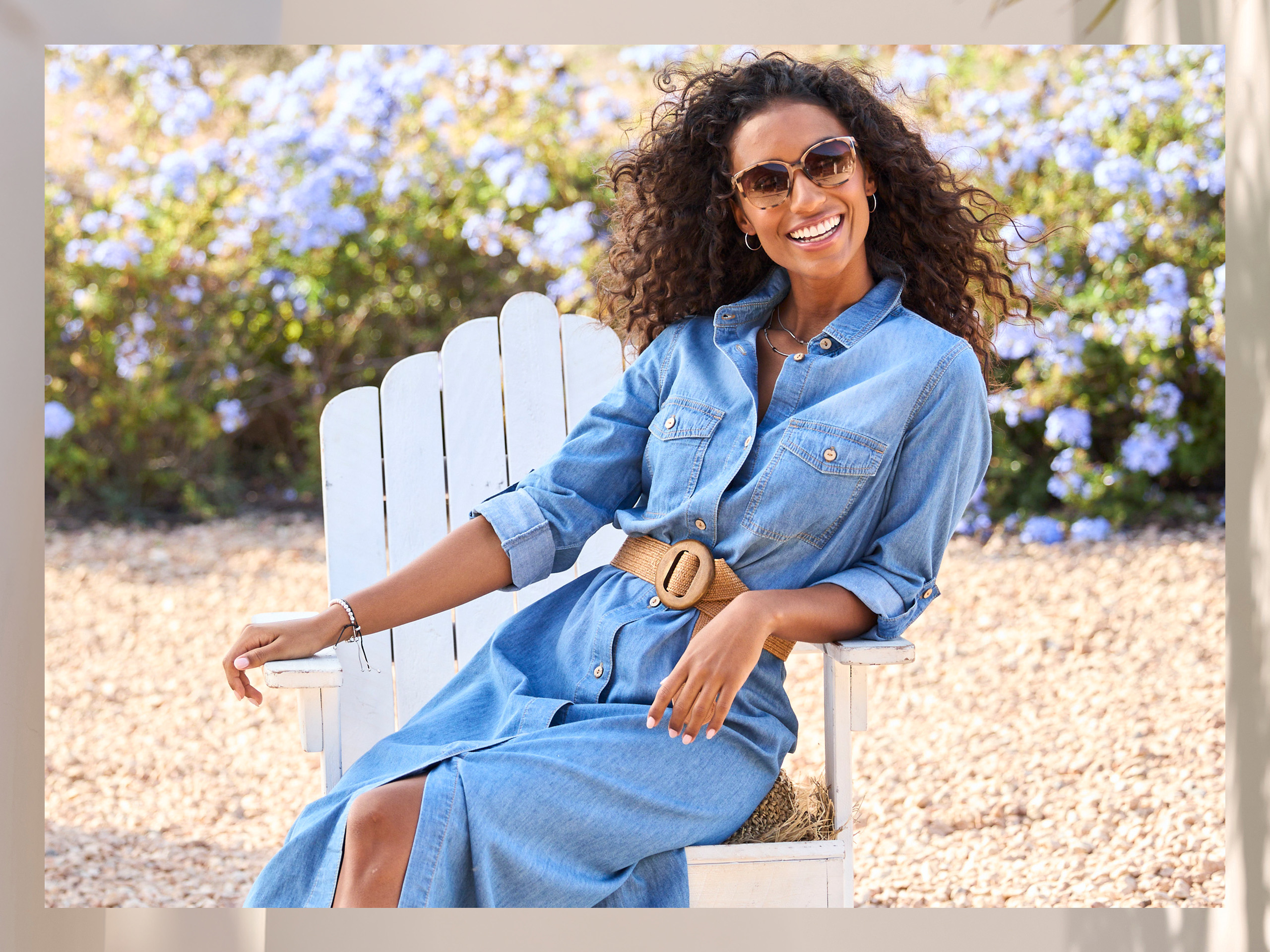 Een model met krullend haar zit op een witte houten stoel, draagt een lange spijkerjurk met riem en heeft de mouwen opgerold.