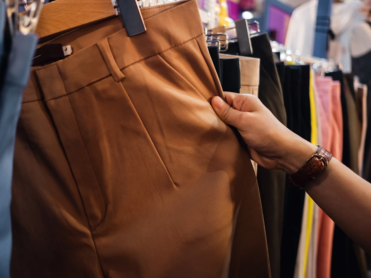 Hand hält eine braune Hose, die an einer Kleiderstange zwischen anderen farbigen Hosen hängt. Hand hält eine braune Hose, die an einer Kleiderstange zwischen anderen farbigen Hosen hängt.
