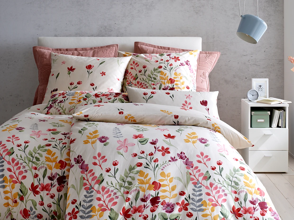 Ein Bett ist mit farbenfroher Bettwäsche im floralen Design bezogen. Im Hintergrund sind eine weiße Nachttischkommode und eine blaue Wandlampe zu sehen.