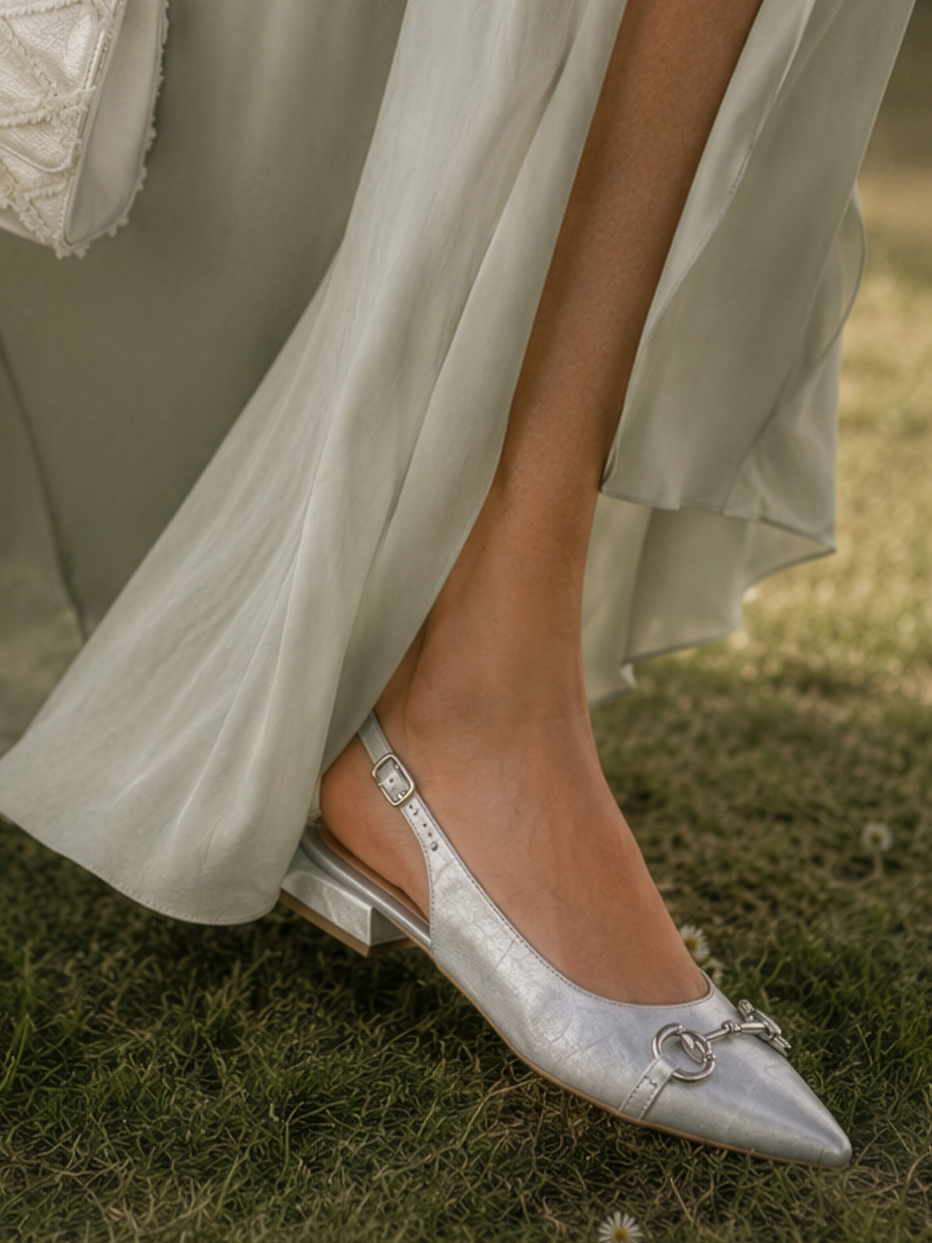 n pied porte une chaussure slingback argentée à petit talon, le mannequin porte une robe fluide vert clair, debout sur l'herbe.