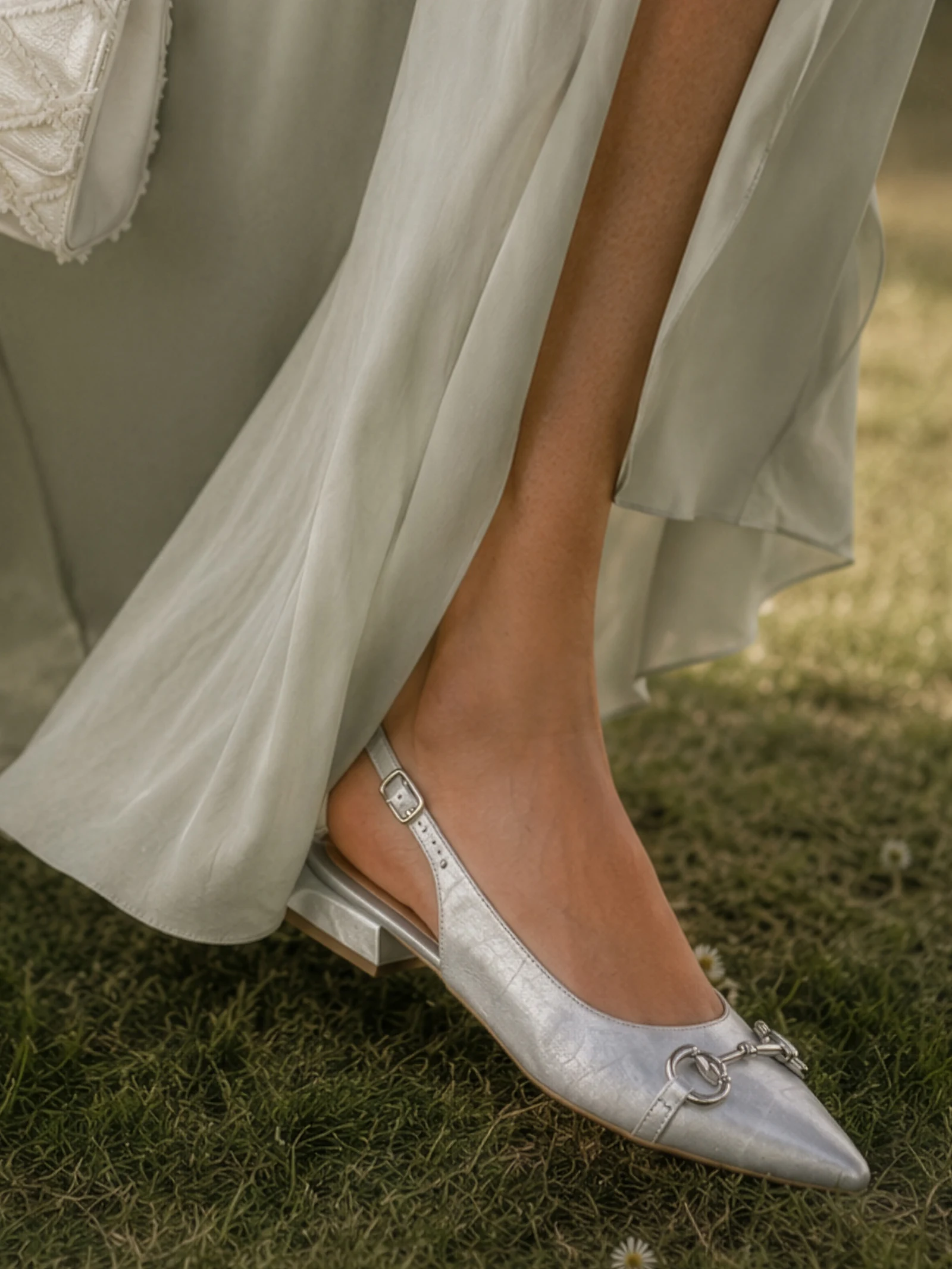 n pied porte une chaussure slingback argentée à petit talon, le mannequin porte une robe fluide vert clair, debout sur l'herbe.