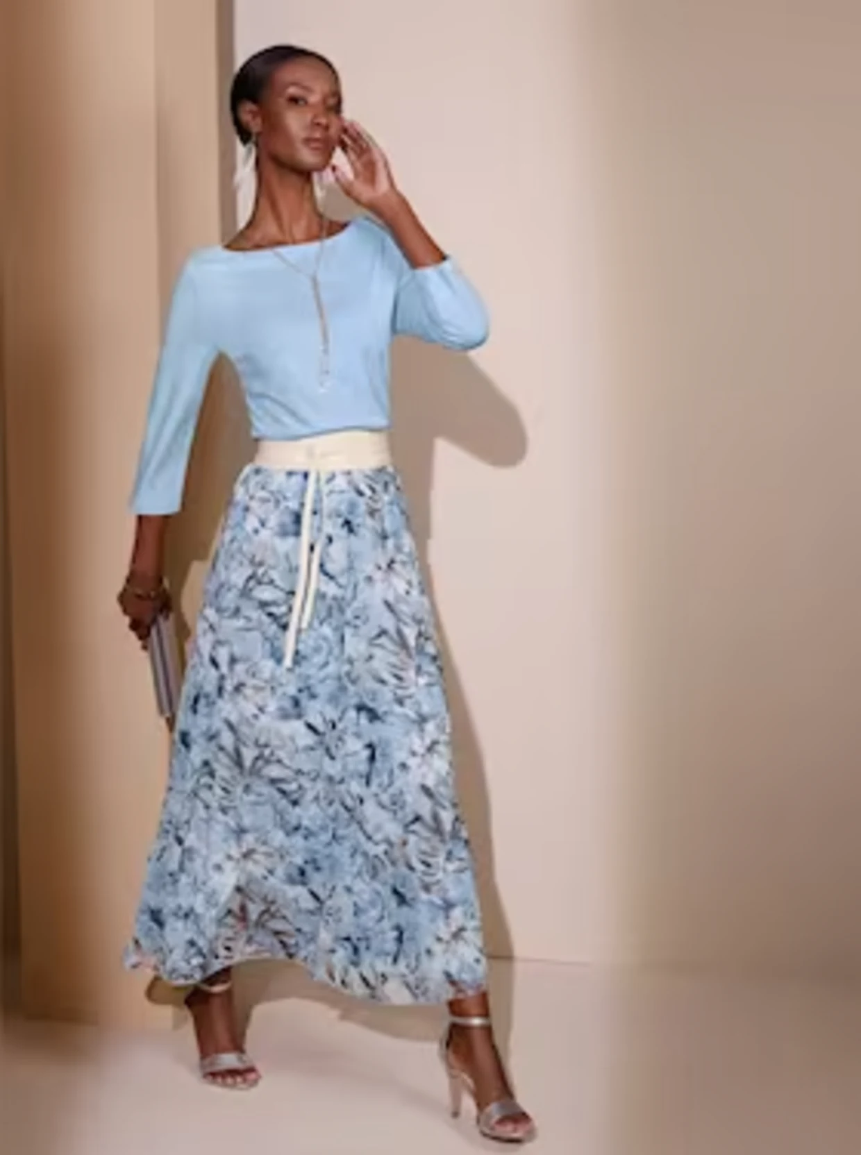 Un modèle porte une élégante jupe bleue à motifs floraux, associée à un haut bleu clair et à une ceinture à la taille. Le look est complété par des sandales hautes et une petite pochette.