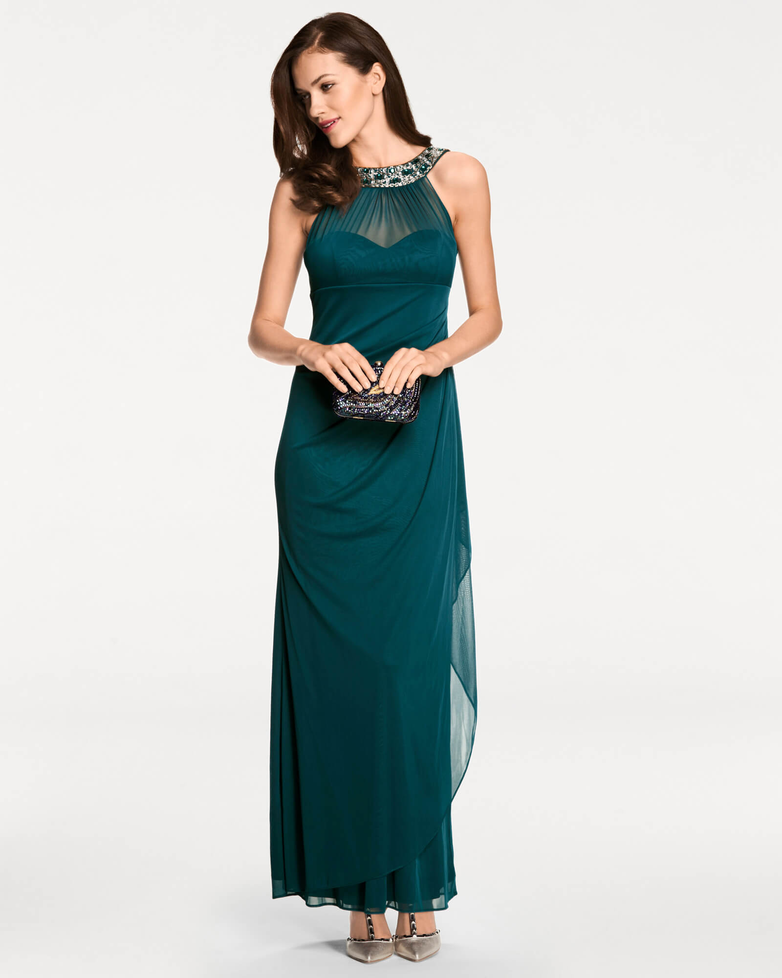 Un mannequin vêtu d'une robe élégante, longue jusqu'au sol, dans un ton vert profond, avec un cou décoratif orné de détails étincelants. La tenue est complétée par une petite pochette brillante.