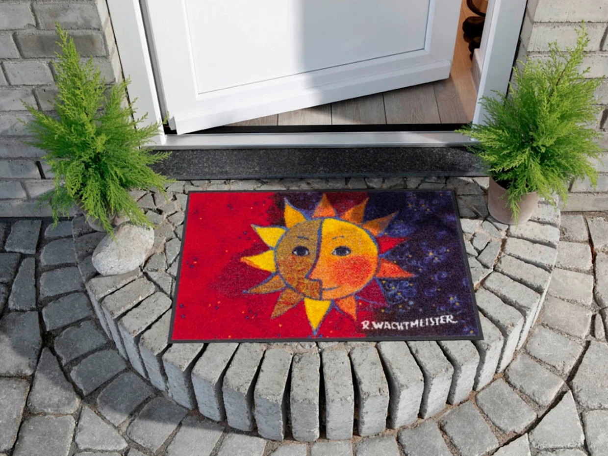 Vor einer weißen Eingangstür liegt eine bunte Fußmatte mit einem stilisierten Sonnenmotiv in Rot, Gelb und Blau. Die Türschwelle ist von zwei grünen Pflanzen in Töpfen eingerahmt, die auf einem gepflasterten Boden stehen.