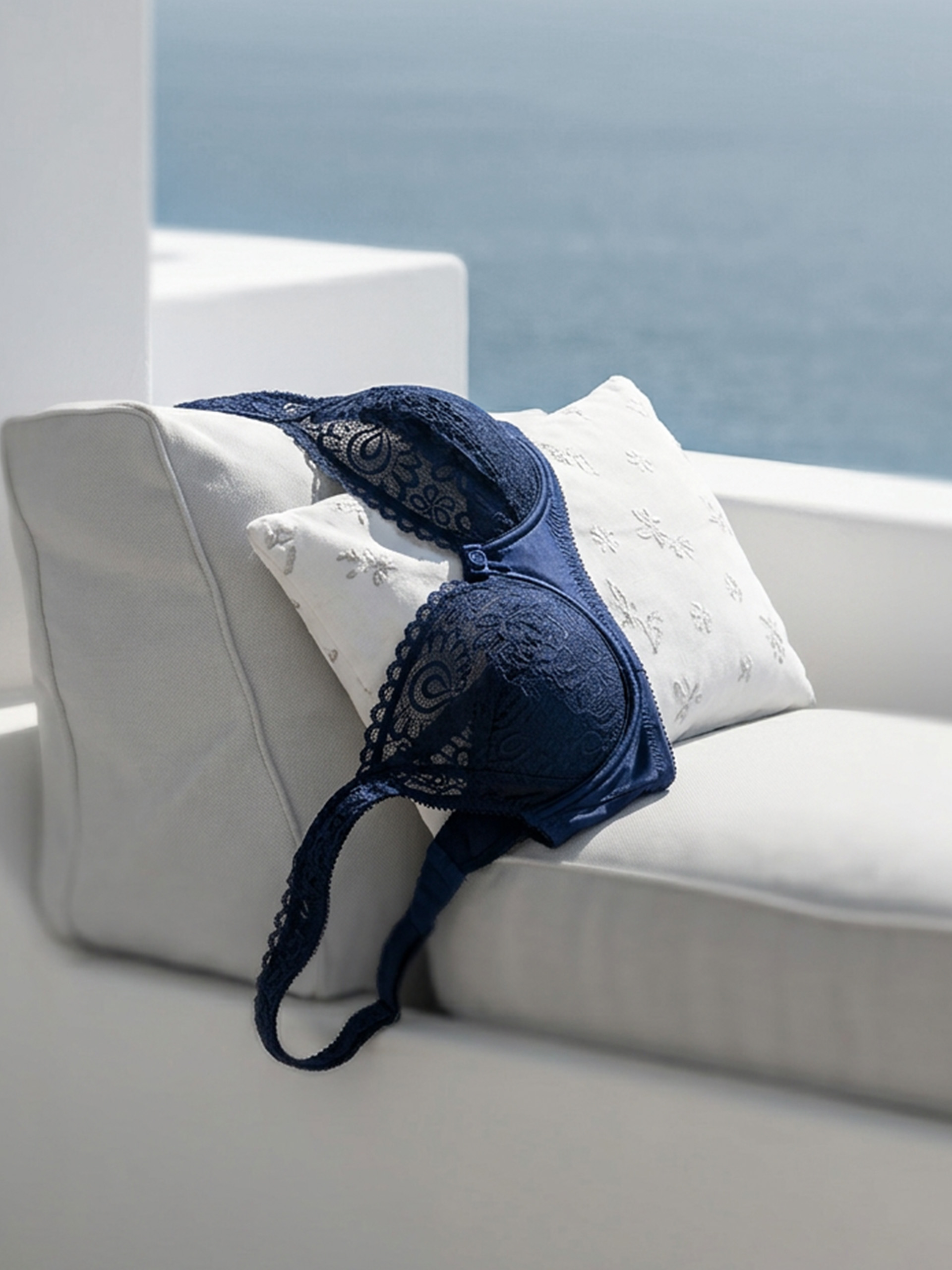 Ein dunkelblauer Spitzen-BH liegt auf einem weißen Kissen auf einem hellen Sofa, im Hintergrund Meer und Himmel.