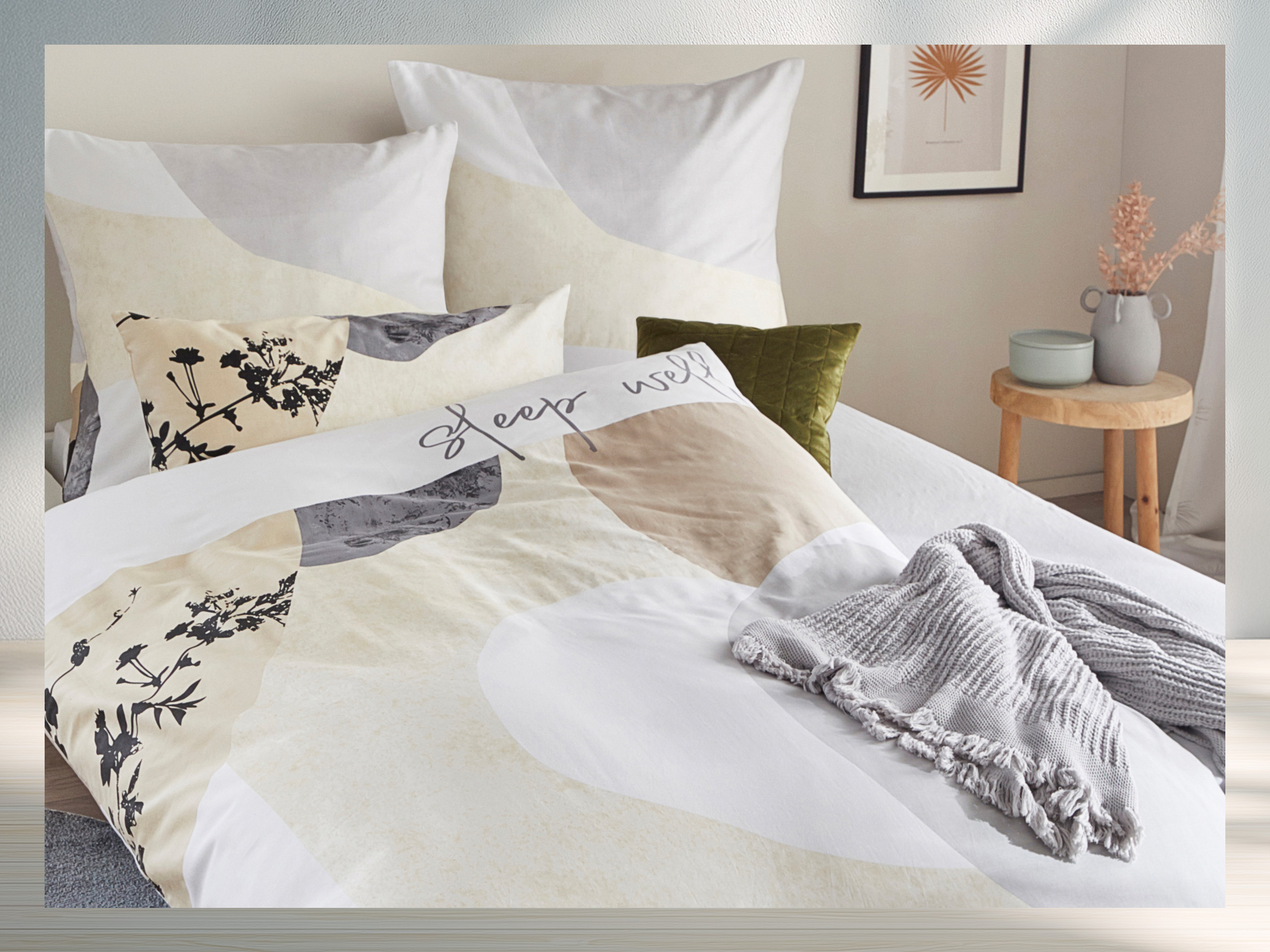 Ein Bett mit hell gemusterter Bettwäsche in Beige, Grau und Weiß, darauf liegt eine graue gestrickte Decke, im Hintergrund Kissen und ein Bild an der Wand.