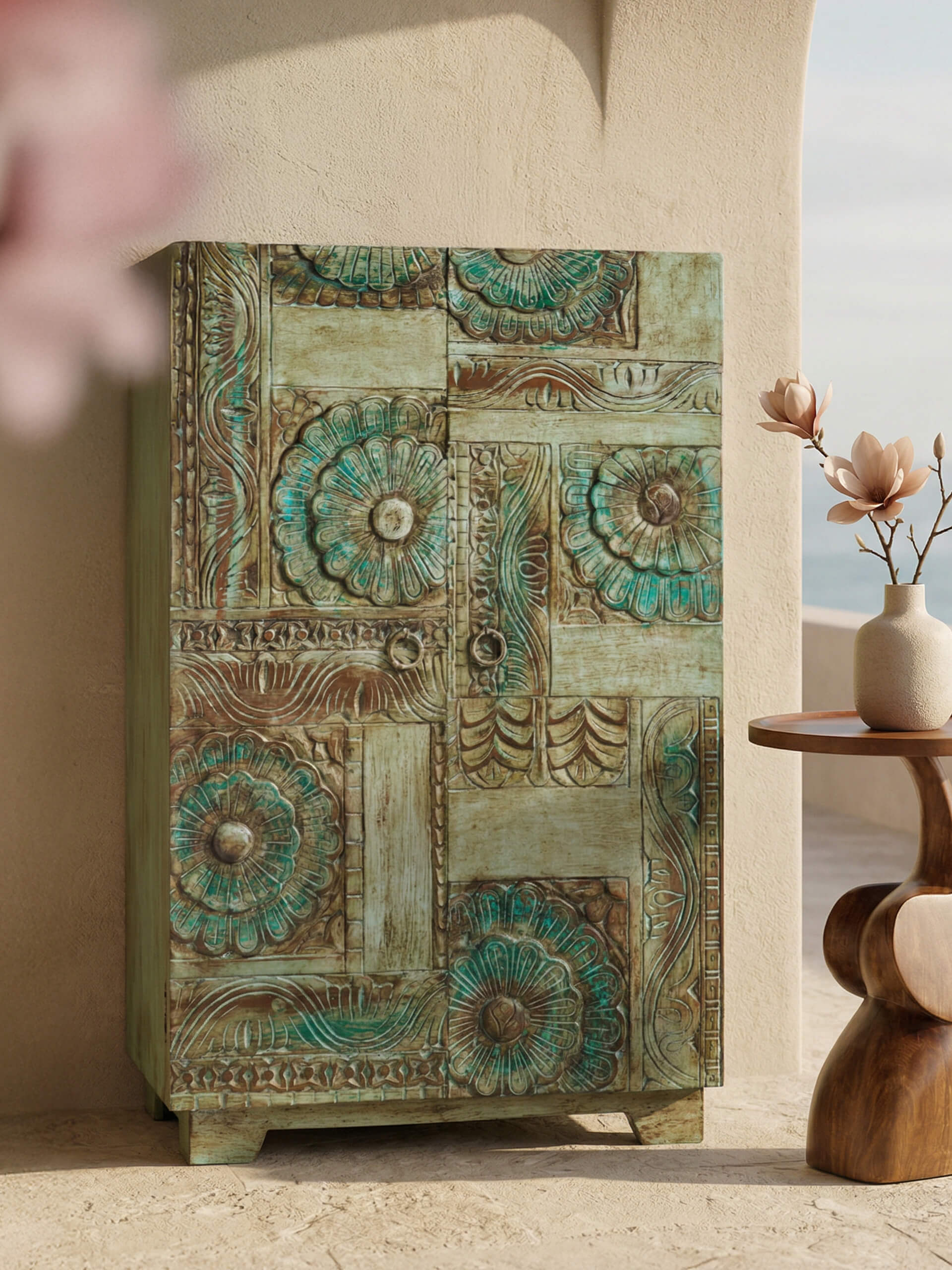 Ein kunstvoll verzierter Schrank mit floralen Mustern in Grün- und Brauntönen steht neben einem kleinen Holztisch mit Vase und Blumen.