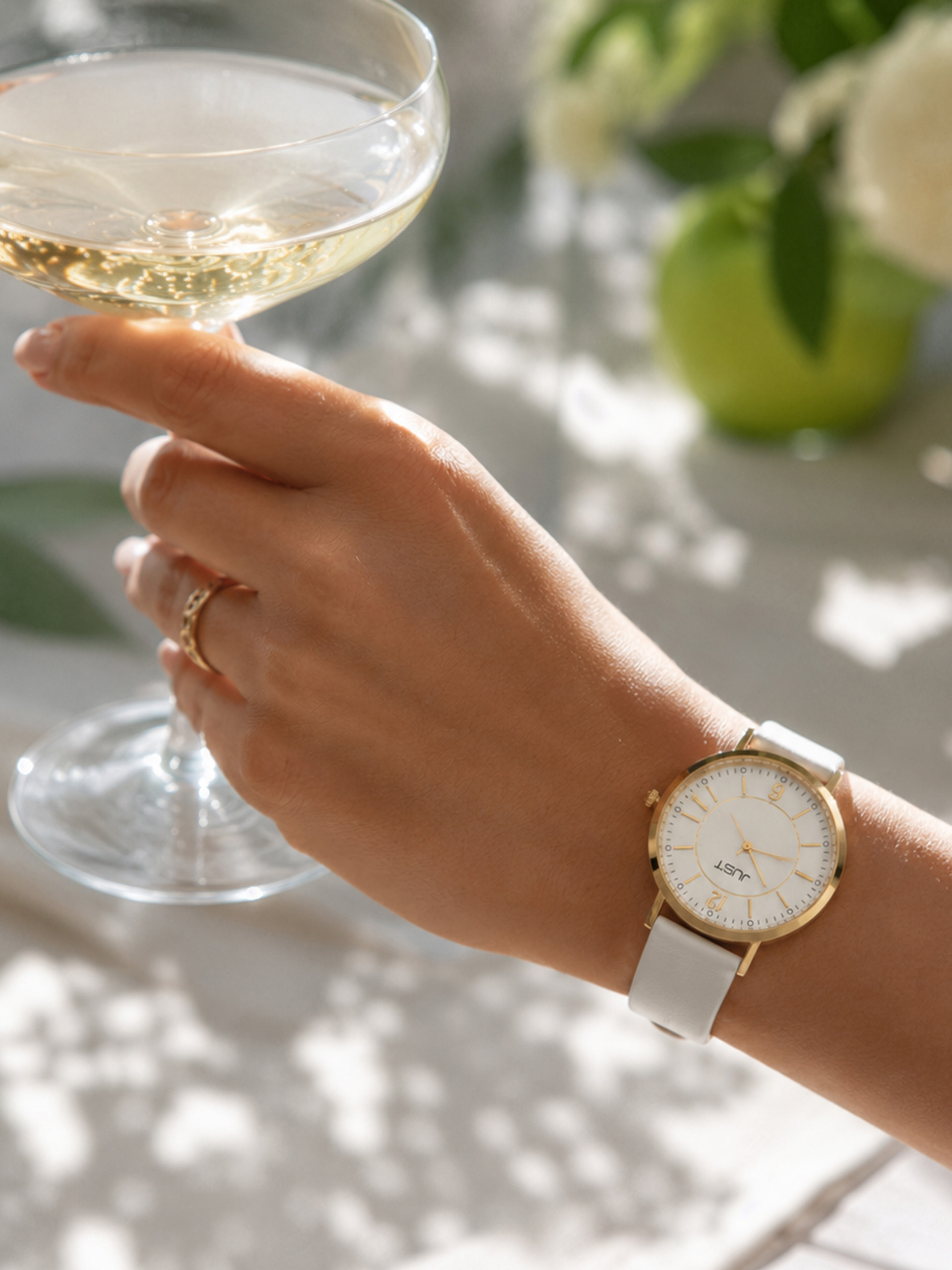 Une main ornée d'une bague dorée et d'un bracelet blanc porte une montre élégante au cadran blanc et tient une coupe de champagne, avec des plantes vertes floues en arrière-plan.