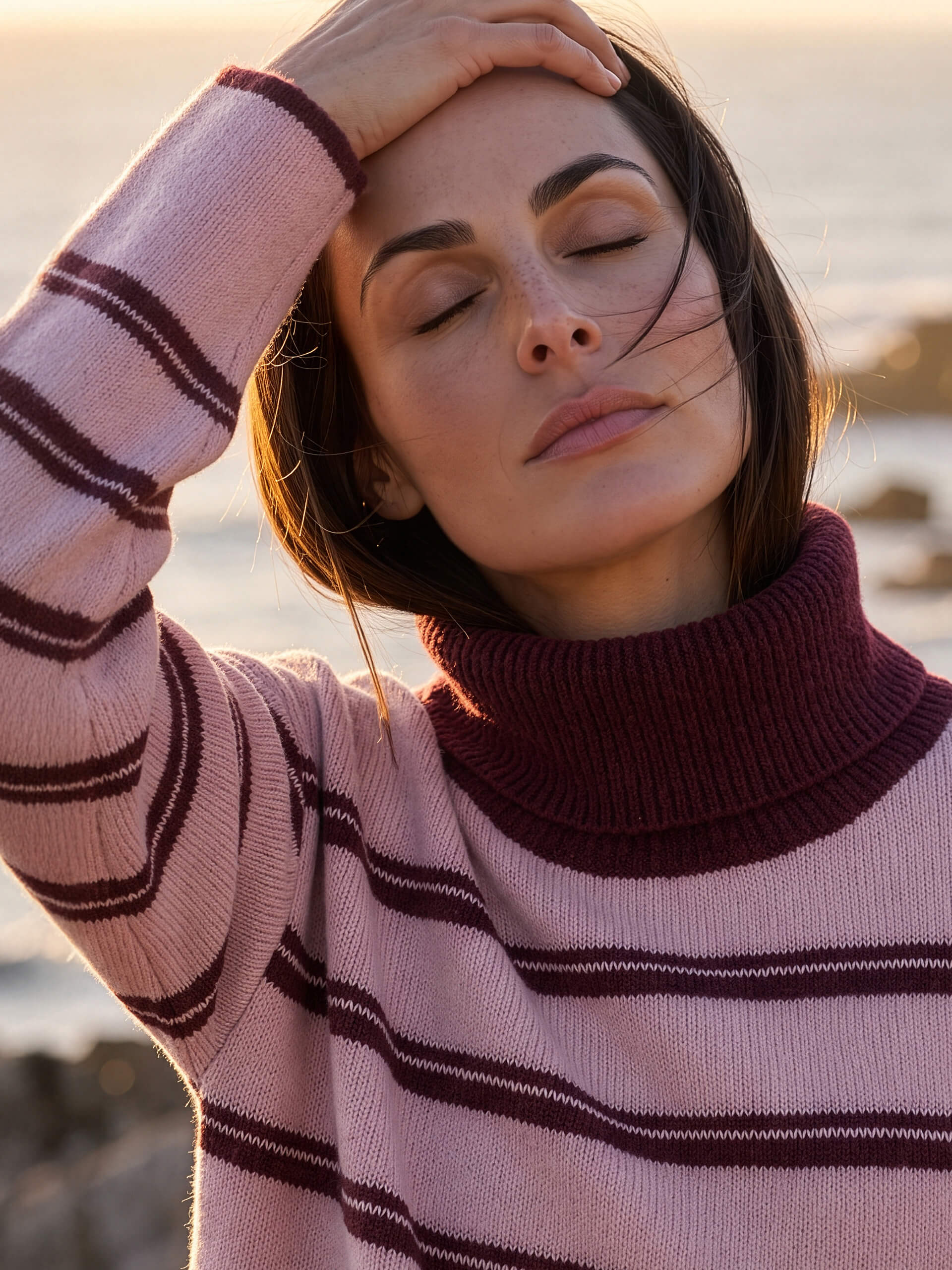 Close-up van een model met gesloten ogen. Ze draagt een roze en wijnrood gestreepte coltrui en houdt een hand op haar voorhoofd.