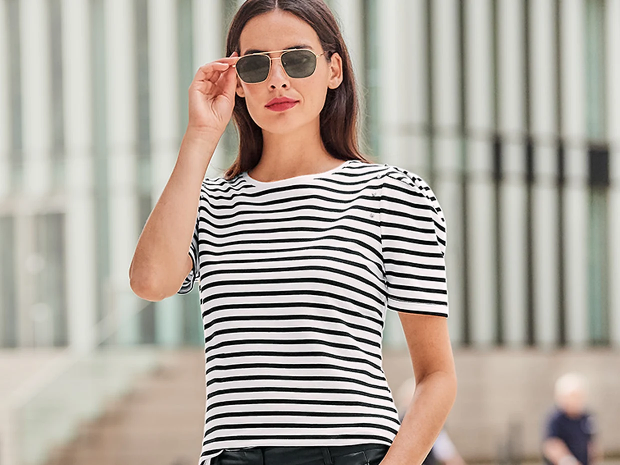 Een model draagt een zwart-wit gestreept shirt terwijl ze buiten staat. De achtergrond toont moderne architectuur met glas en beton.