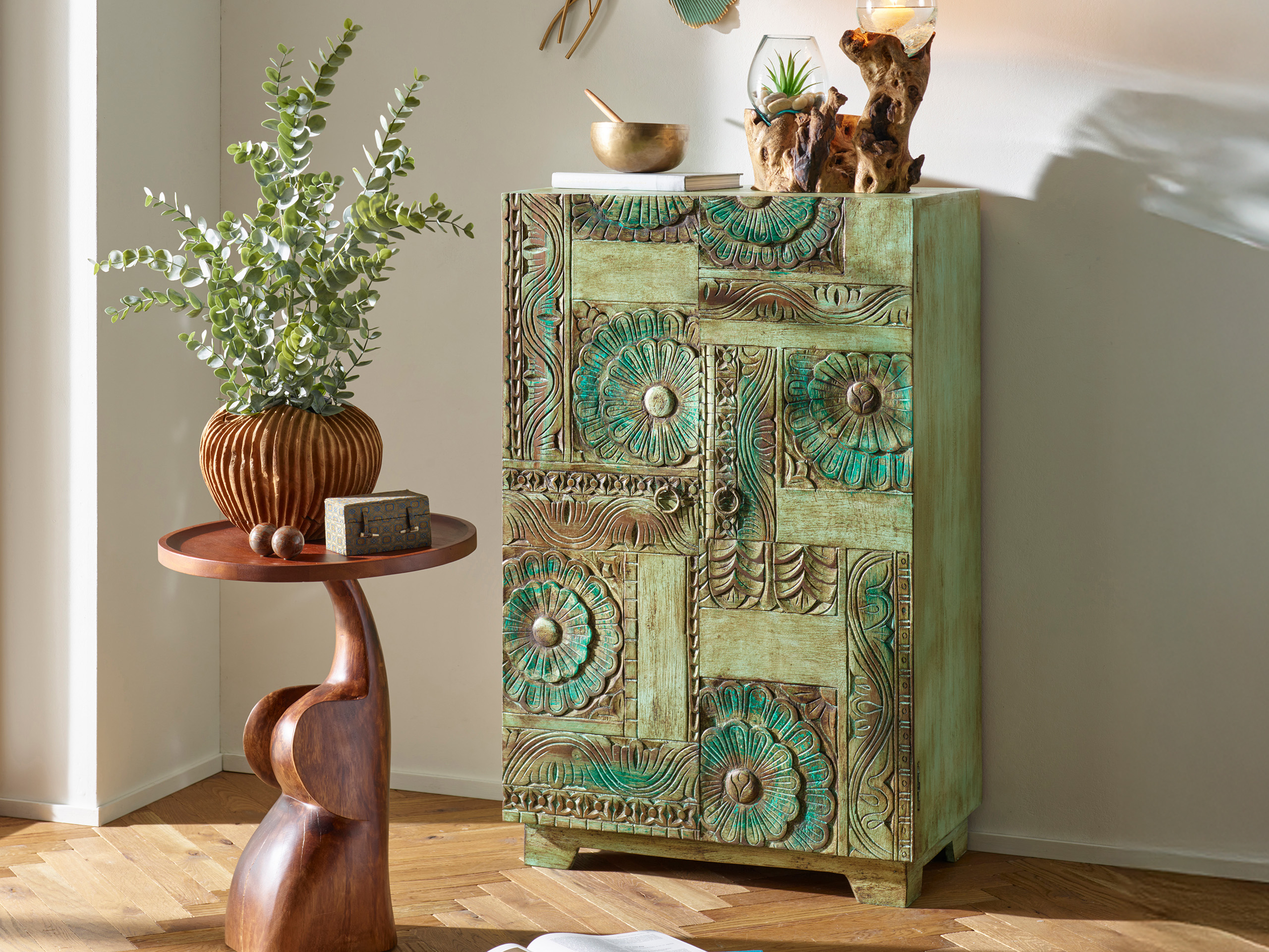 Een groene, kunstig versierde kast staat naast een kleine, bruine bijzettafel met een vaas en planten, in een lichte kamer met raam.