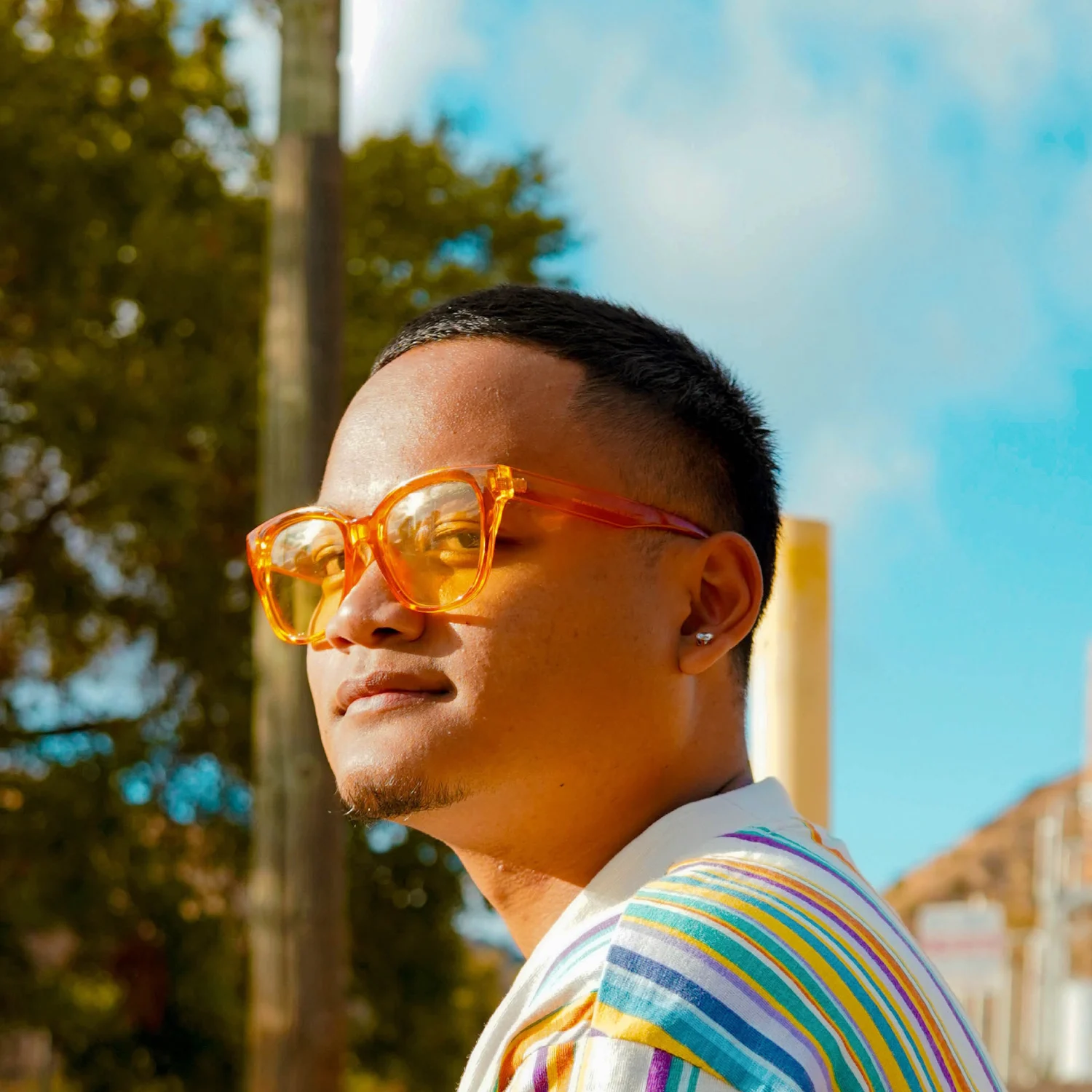 Close-up of a person with short hair wearing orange-tinted sunglasses, looking to the side with a clear blue sky in the background. Individual is wearing a colorful shirt on a sunny outdoor setting.