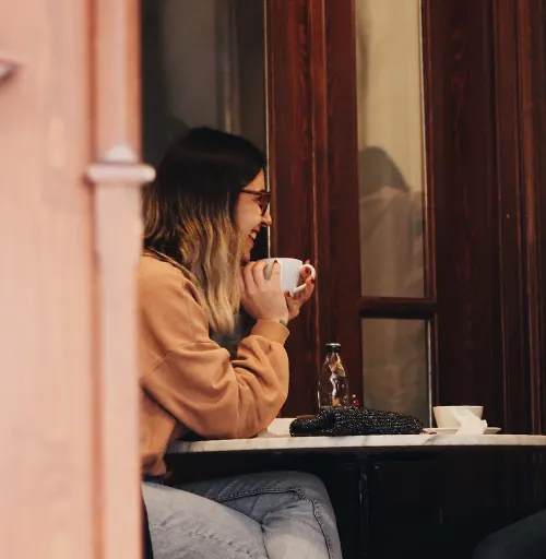 A smiling woman in glasses holding a coffee cup, seated at a table with a beaded purse and a bottle, in a cozy cafe setting along with the words 'budgeting for vacation' on top of the image.