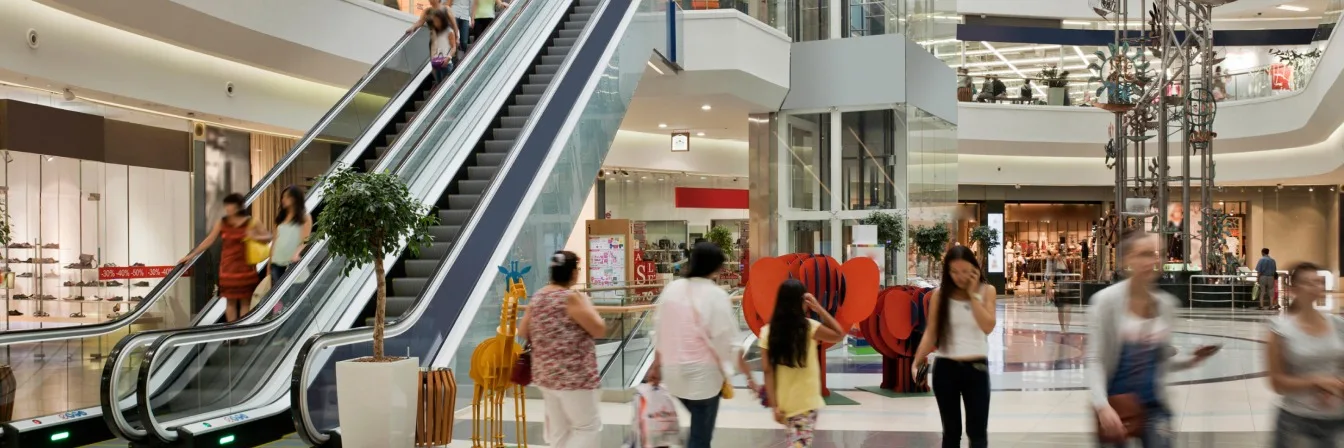Modern shopping mall interior with escalators, glass elevator, and shoppers moving between multiple levels of retail stores.