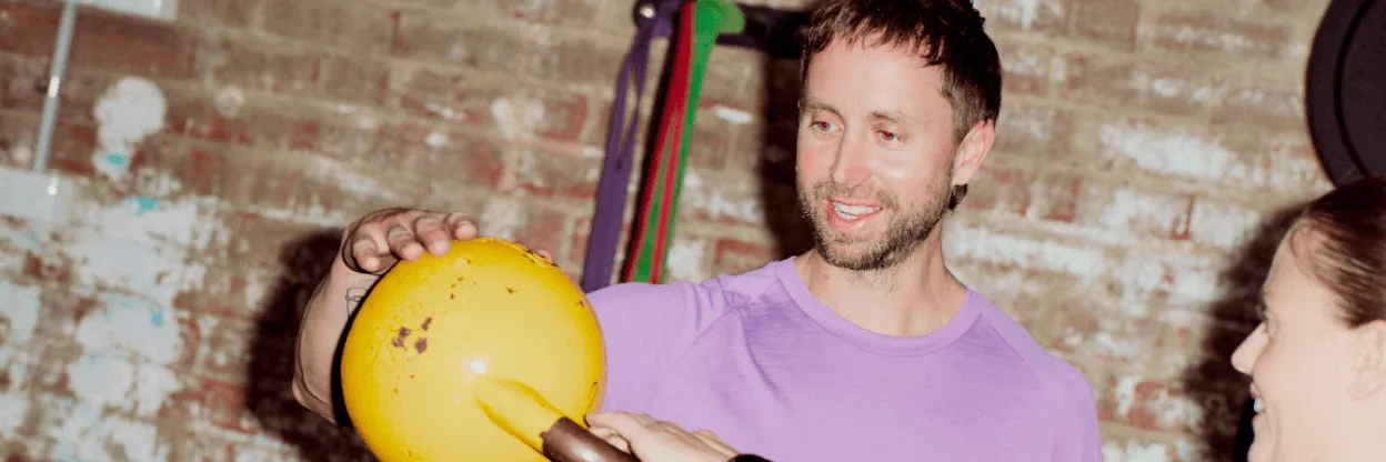 Person in purple shirt holding a yellow exercise ball against brick wall background with colorful resistance bands.