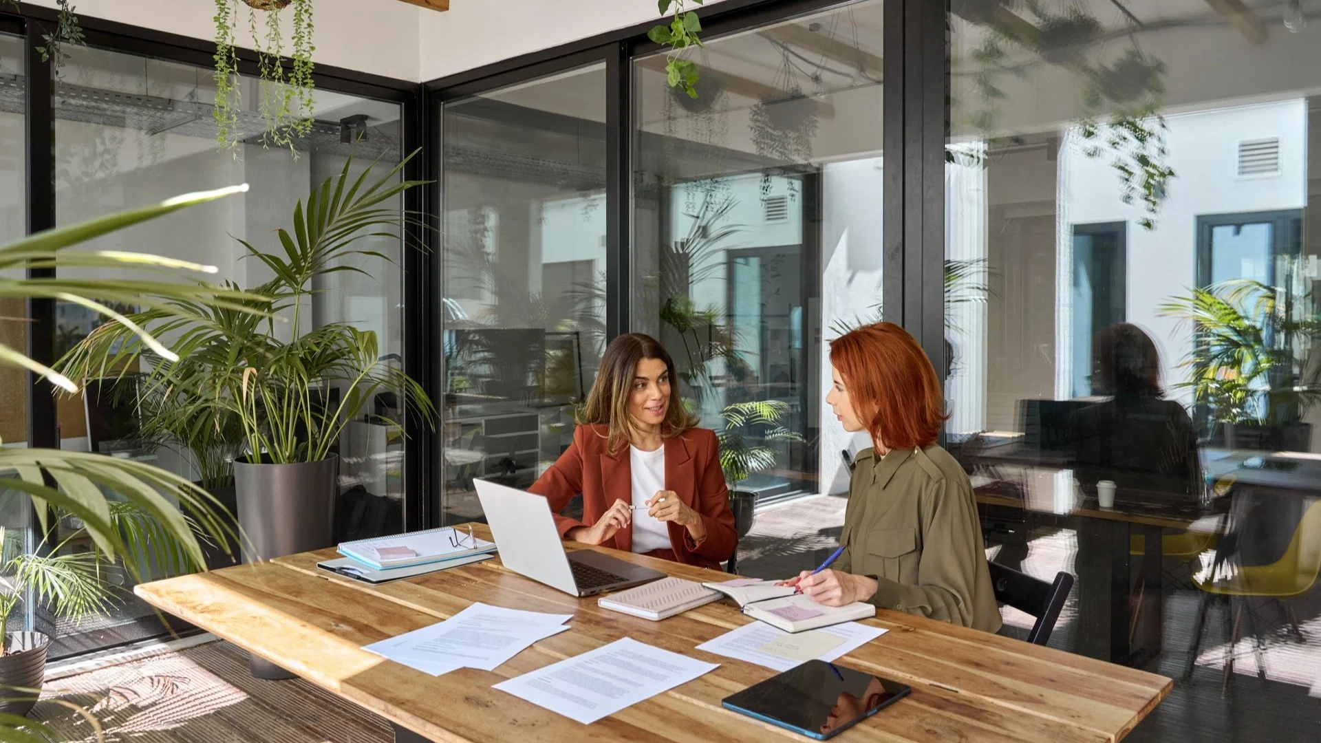 Two women in business meeting at wooden table with laptop and documents in modern office surrounded by plants.