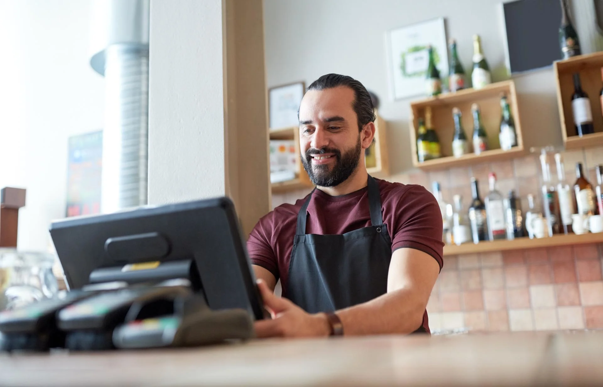 Smiling bartender in burgundy shirt and black apron using a touchscreen register with bottles displayed on shelves behind.