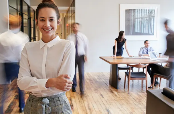 A business woman stands in a modern office, arms folded, smiling. In the background are people walking about, working at desks and in conversation.