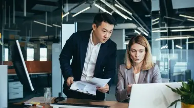 Two professionals in a modern office reviewing documents together, one standing and one seated at a laptop.