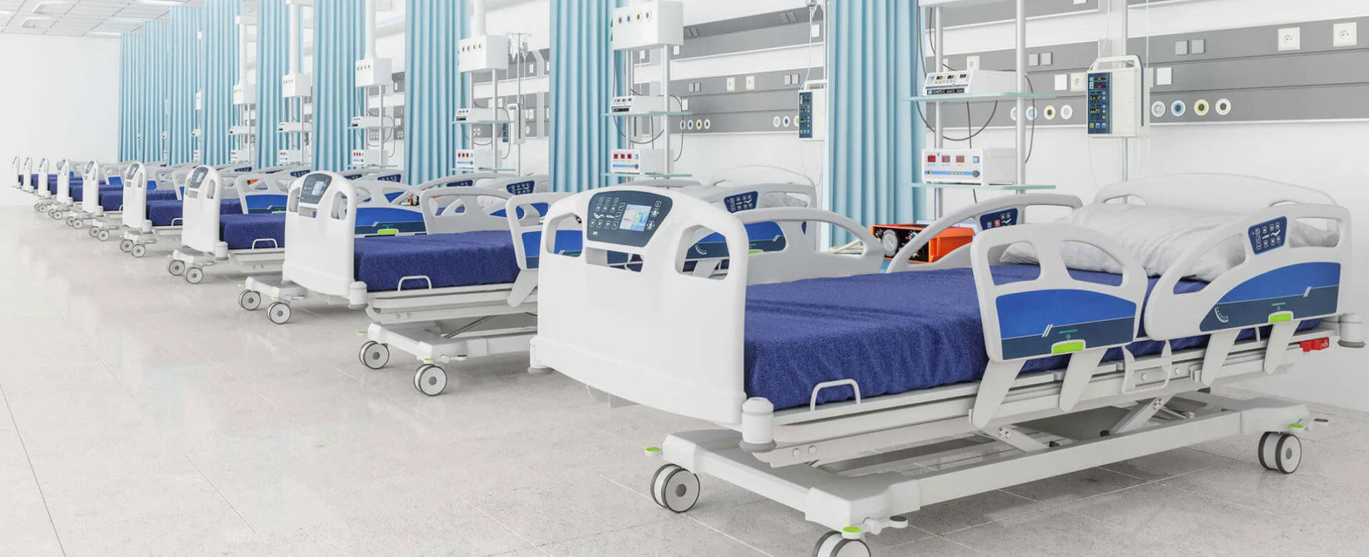 Hospital ward with empty beds with blue covers, separated by light blue privacy curtains and medical monitoring equipment.