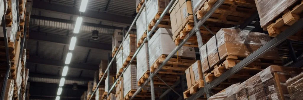 Large warehouse interior with tall metal shelving stacked with cardboard boxes and pallets under fluorescent lighting.