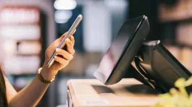 Person using smartphone next to a point-of-sale terminal at a retail counter in a dimly lit store.